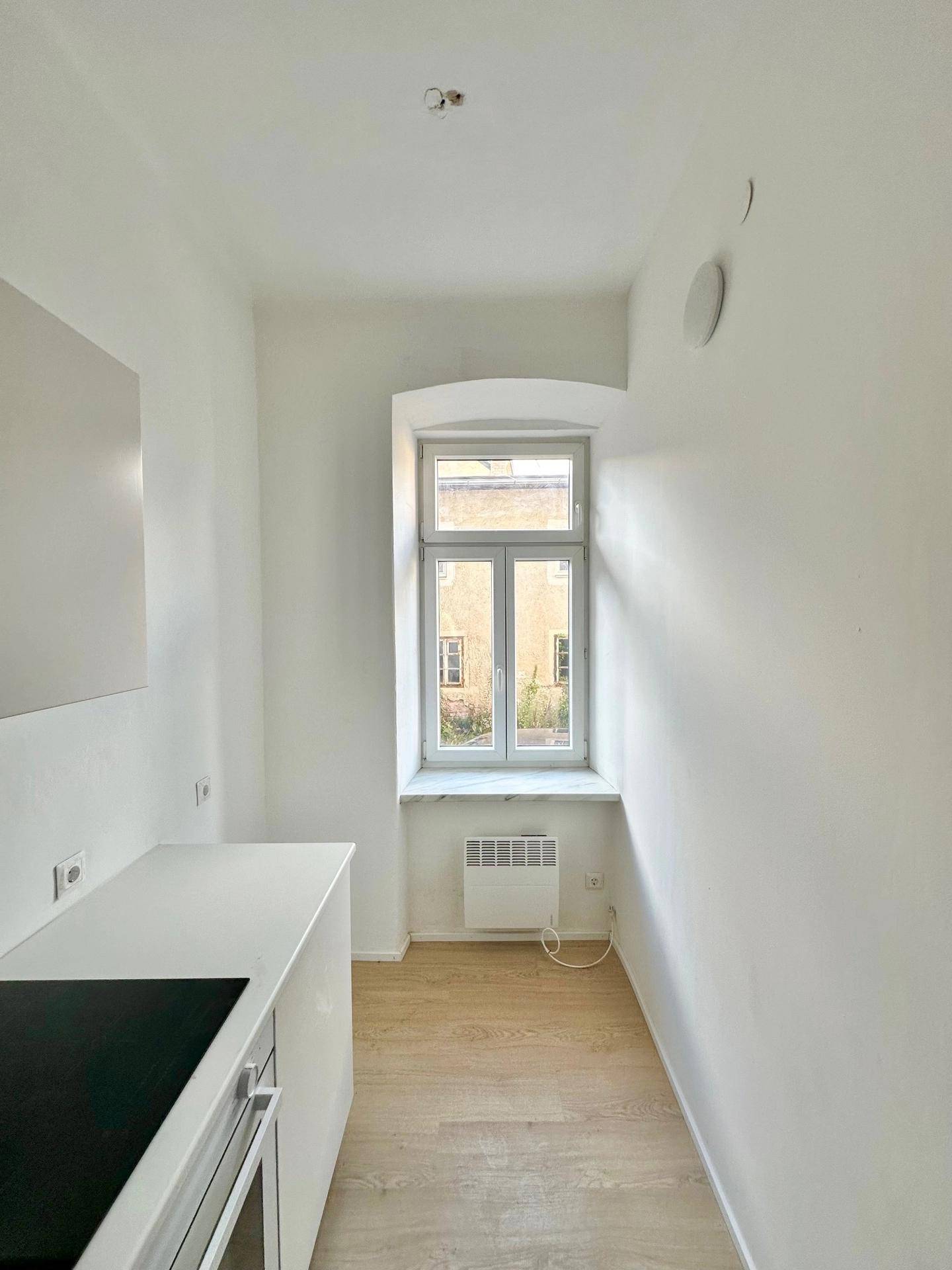 Schmale Küche mit hellem Holzboden, weißen Schränken und Fenster.
