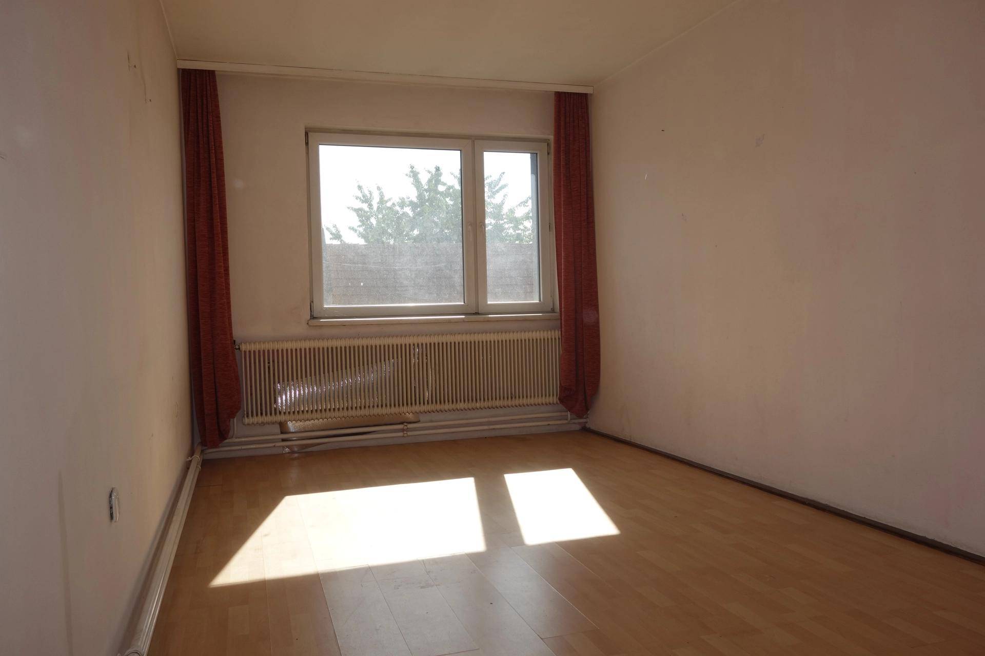Leeres Zimmer mit hellem Laminatboden, roten Vorhängen und viel natürlichem Licht.