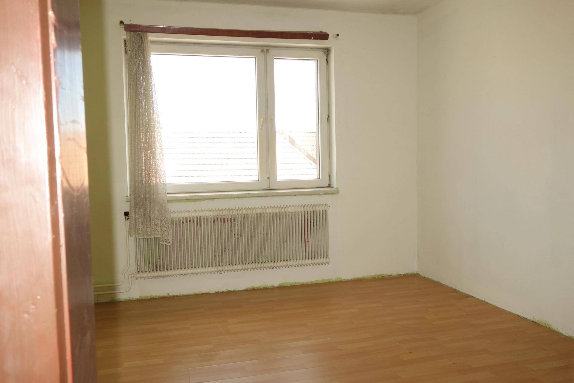 Helles, leeres Zimmer mit weißen Wänden, Laminatboden, Fenster und Heizkörper.