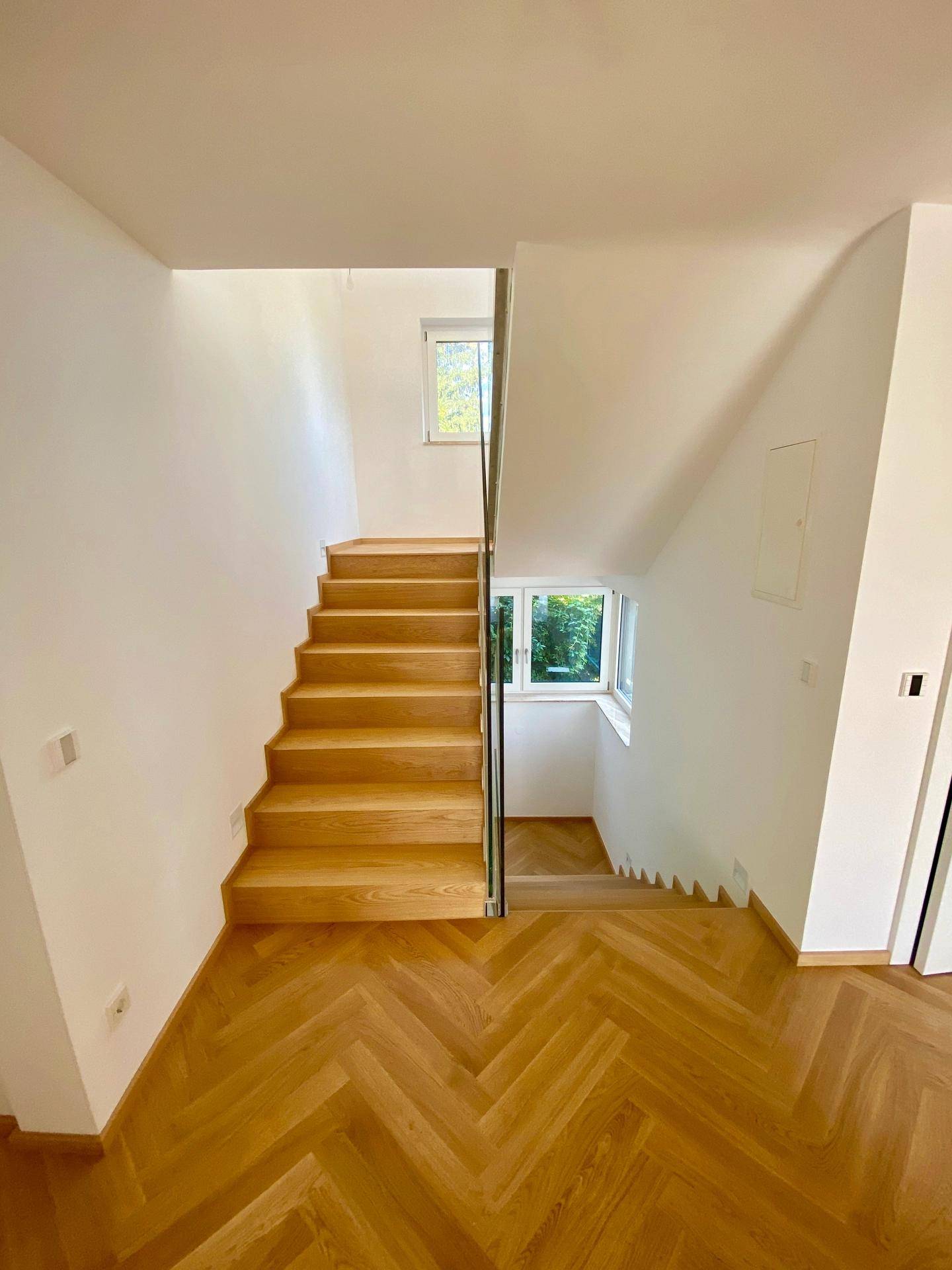 Heller Treppenaufgang mit Holzstufen, Glasgeländer und Fischgrätparkett.
