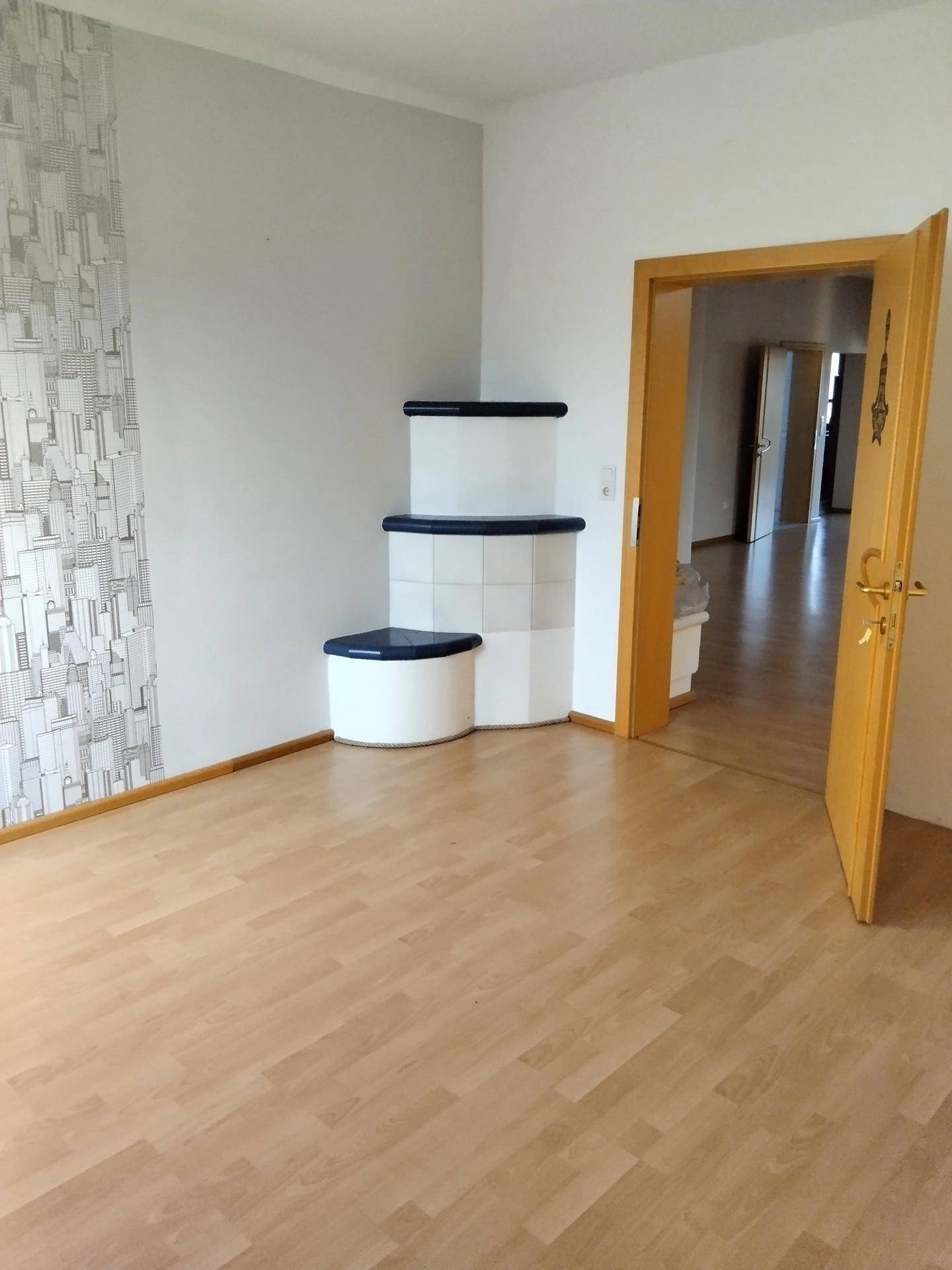 Geräumiges Zimmer mit hellem Laminatboden und einem traditionellen Kachelofen in der Ecke.