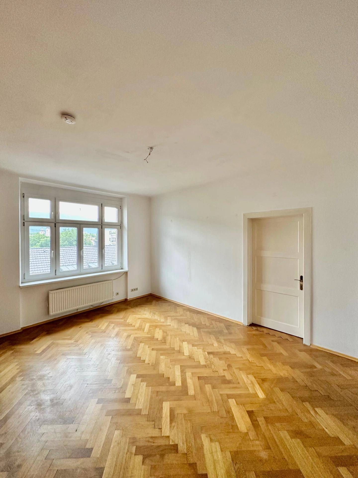 Helles Zimmer mit Fischgrätparkett, weißer Tür und Blick auf Dächer und Bäume.