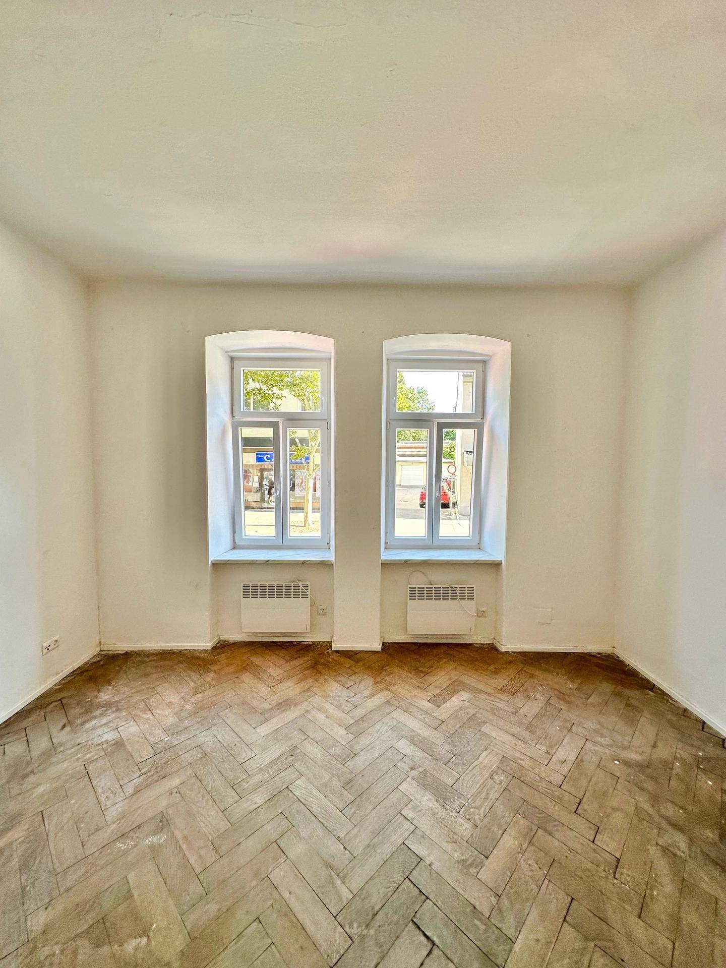 Geräumiger Raum mit Fischgrätparkett und zwei großen Fenstern zur Straße.