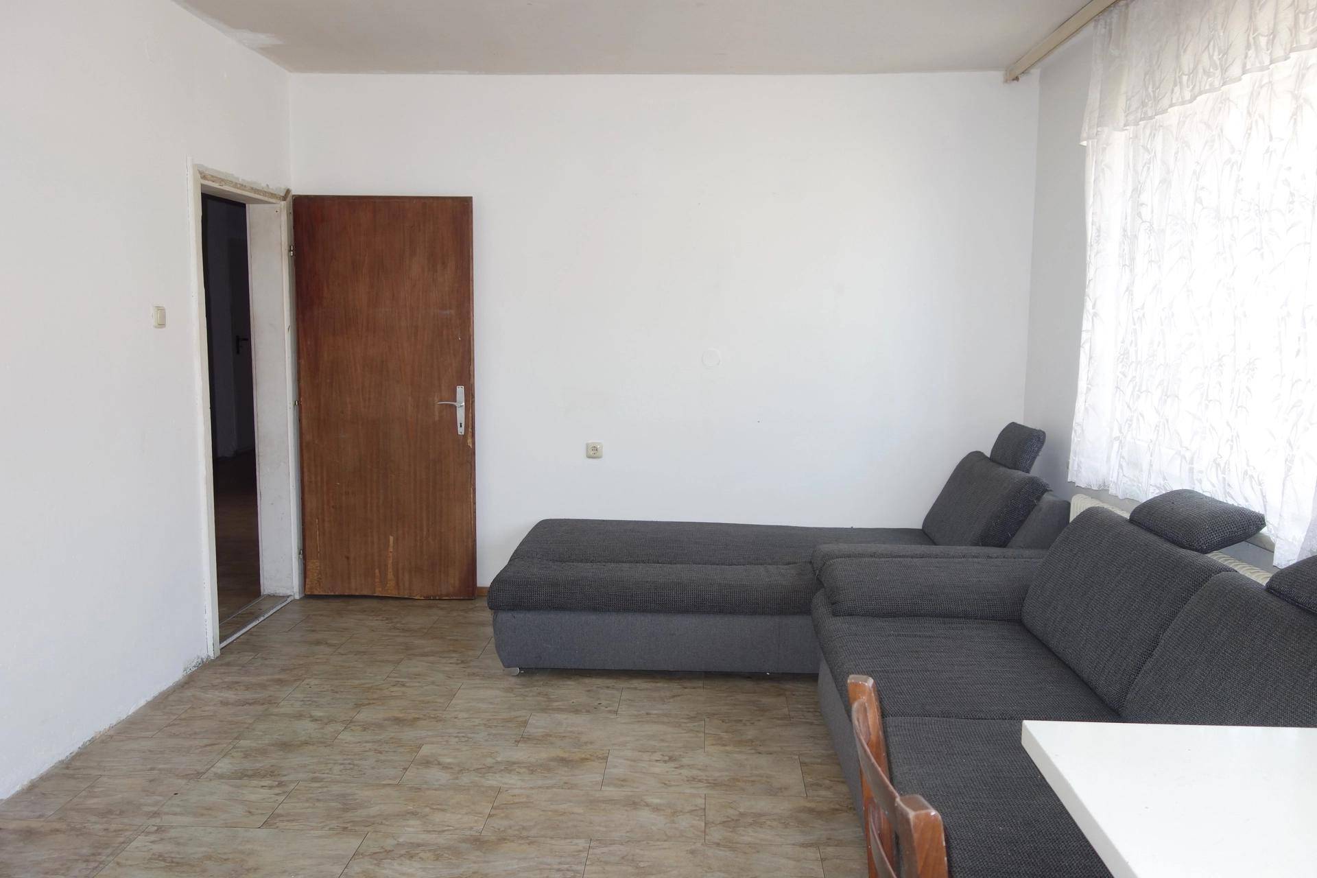 Wohnzimmer mit großem grauen Ecksofa, gefliestem Boden und einem Fenster mit Vorhängen.