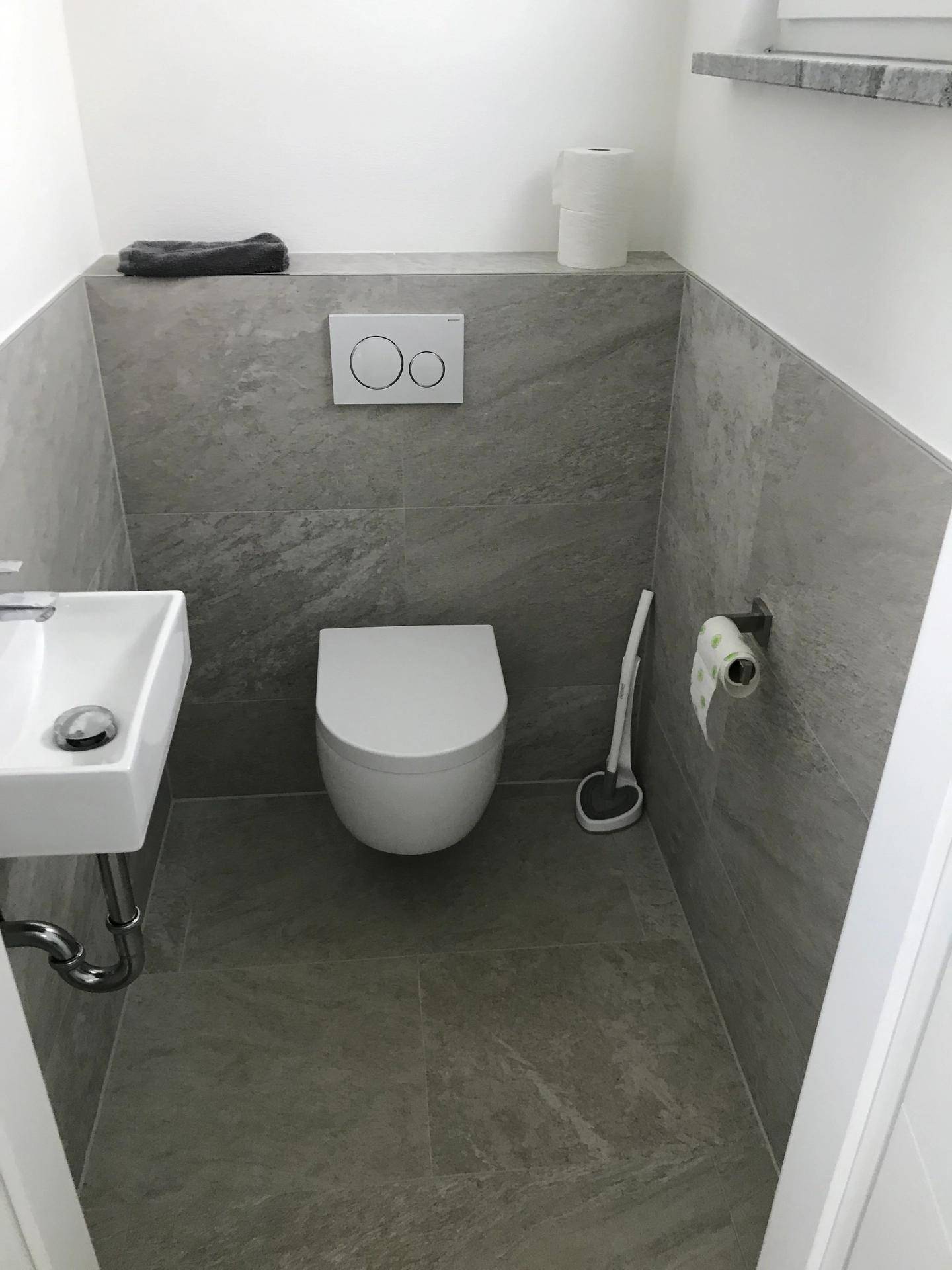 Modernes Gäste-WC mit wandhängender Toilette, kleinem Waschbecken und grauen Fliesen.