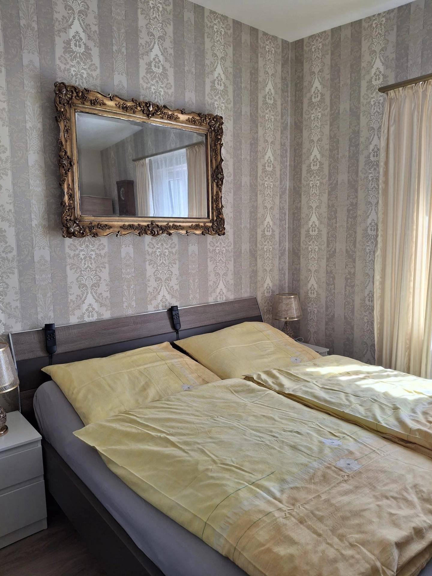 Schlafzimmer mit gemusterter Tapete, Doppelbett und einem großen, verzierten Spiegel.