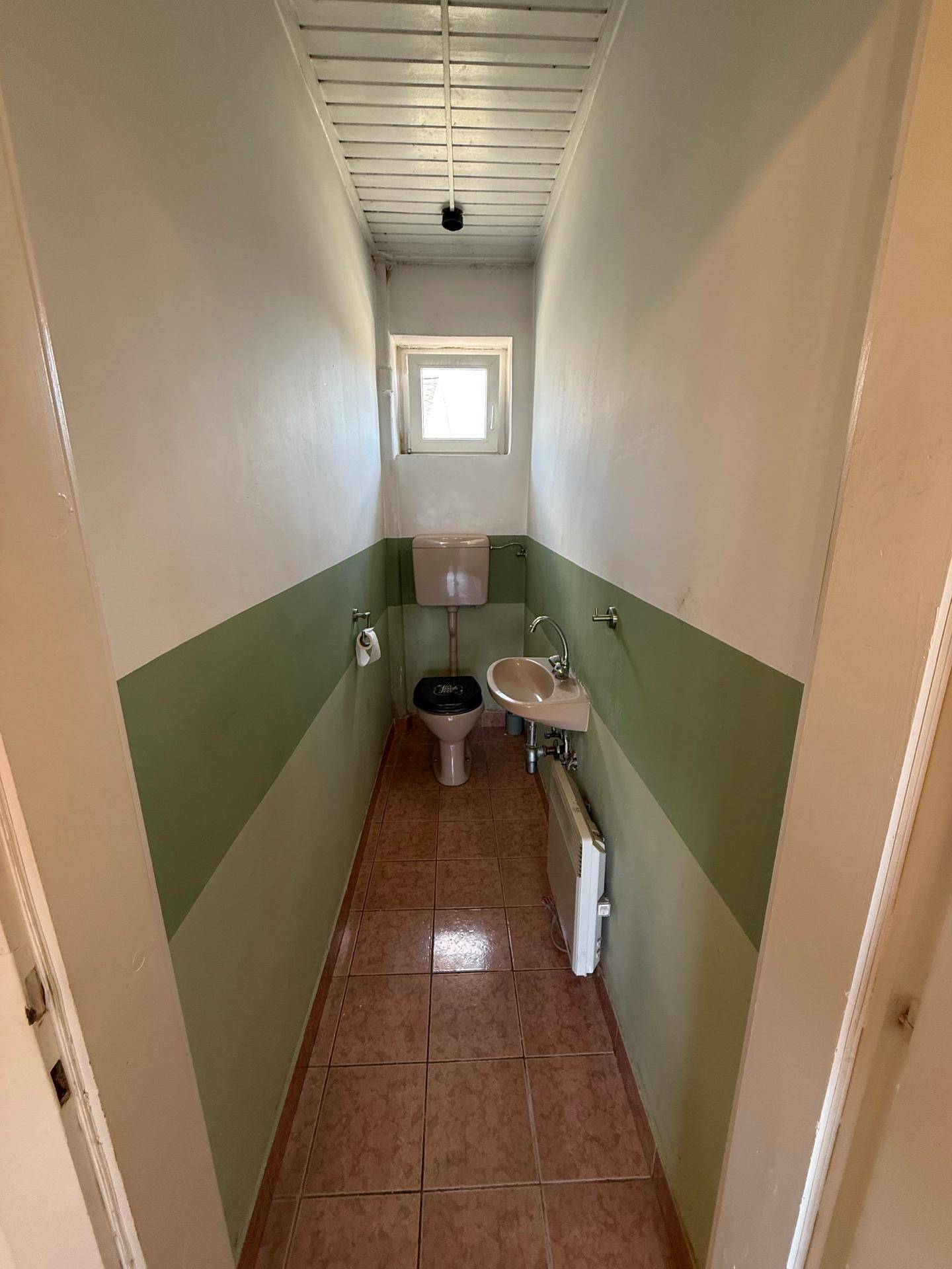 Kleines WC mit Fliesenboden, Toilette, Waschbecken und einem Fenster für Tageslicht.