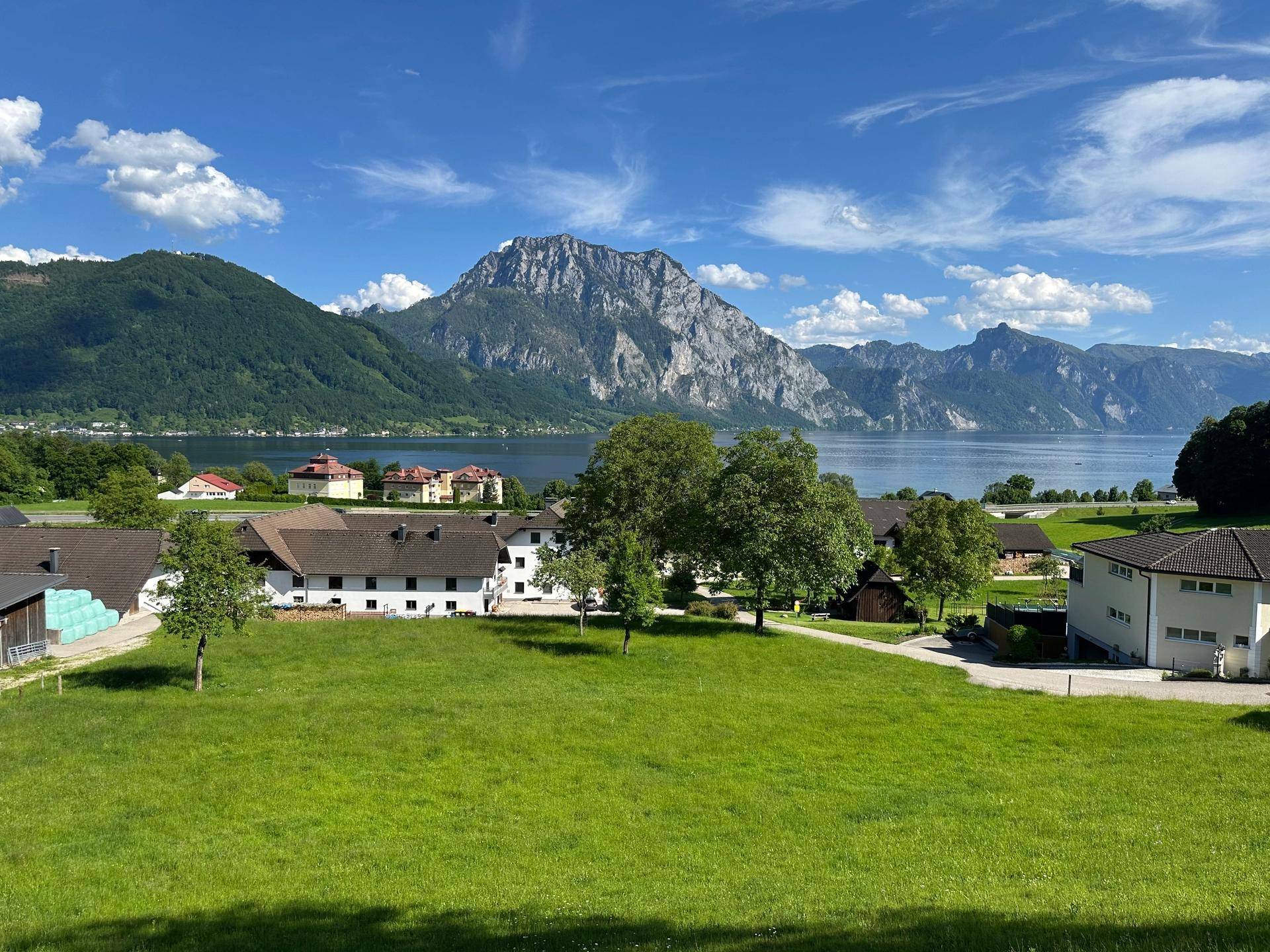 Weitläufige grüne Wiesen mit Blick auf den See und die majestätischen Berge unter blauem Himmel.
