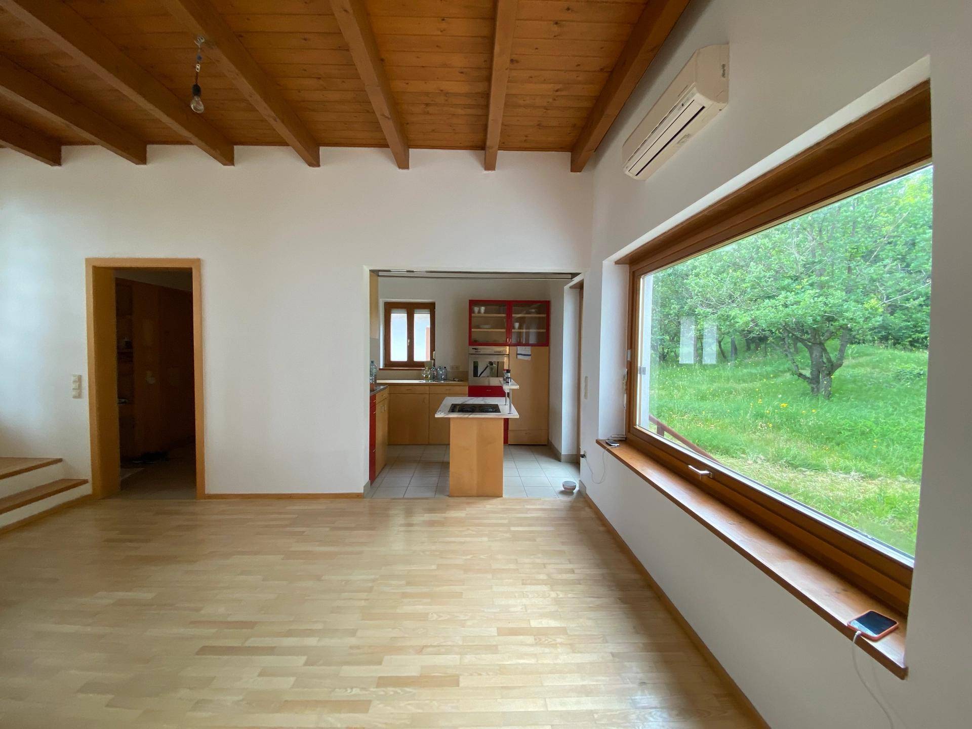 Geräumiges Wohnzimmer mit Holzboden, freiliegenden Deckenbalken und Blick ins Grüne.