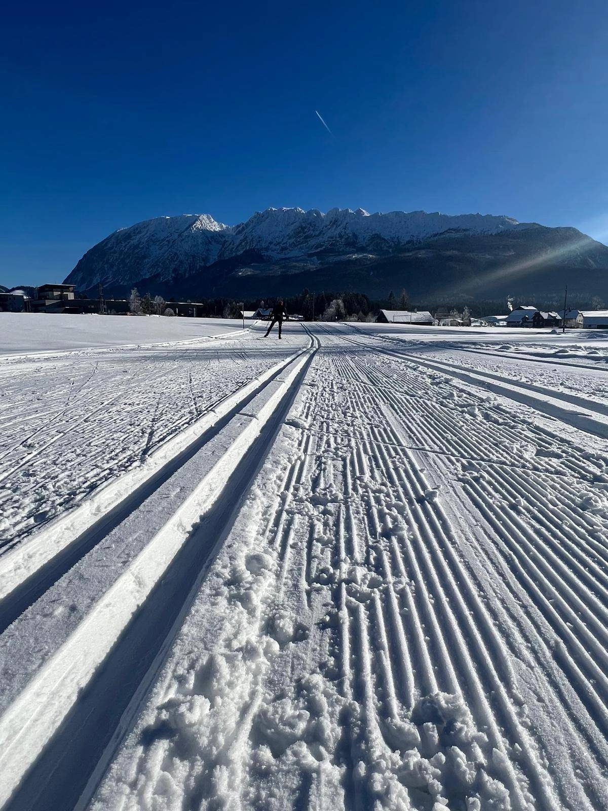 Gespurtete Langlaufloipe in einer weiten Winterlandschaft mit schneebedeckten Bergen im Hintergrund.