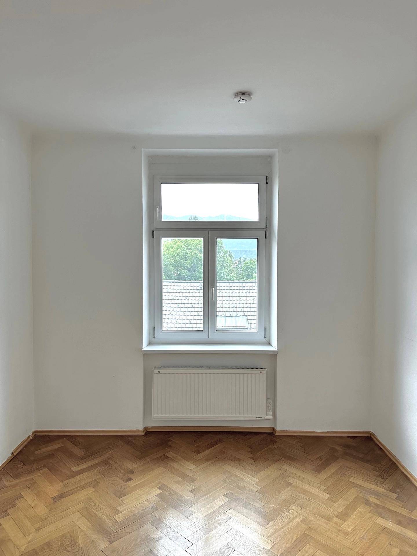 Leeres Zimmer mit Fischgrätparkett, Heizkörper und Blick auf Dächer und Bäume.