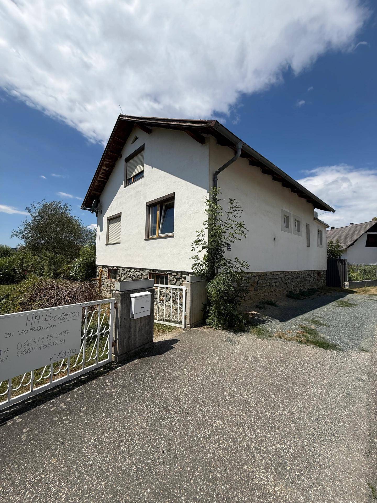Freistehendes Haus mit weißer Fassade, Steinsockel und Einfahrt unter sonnigem Himmel.