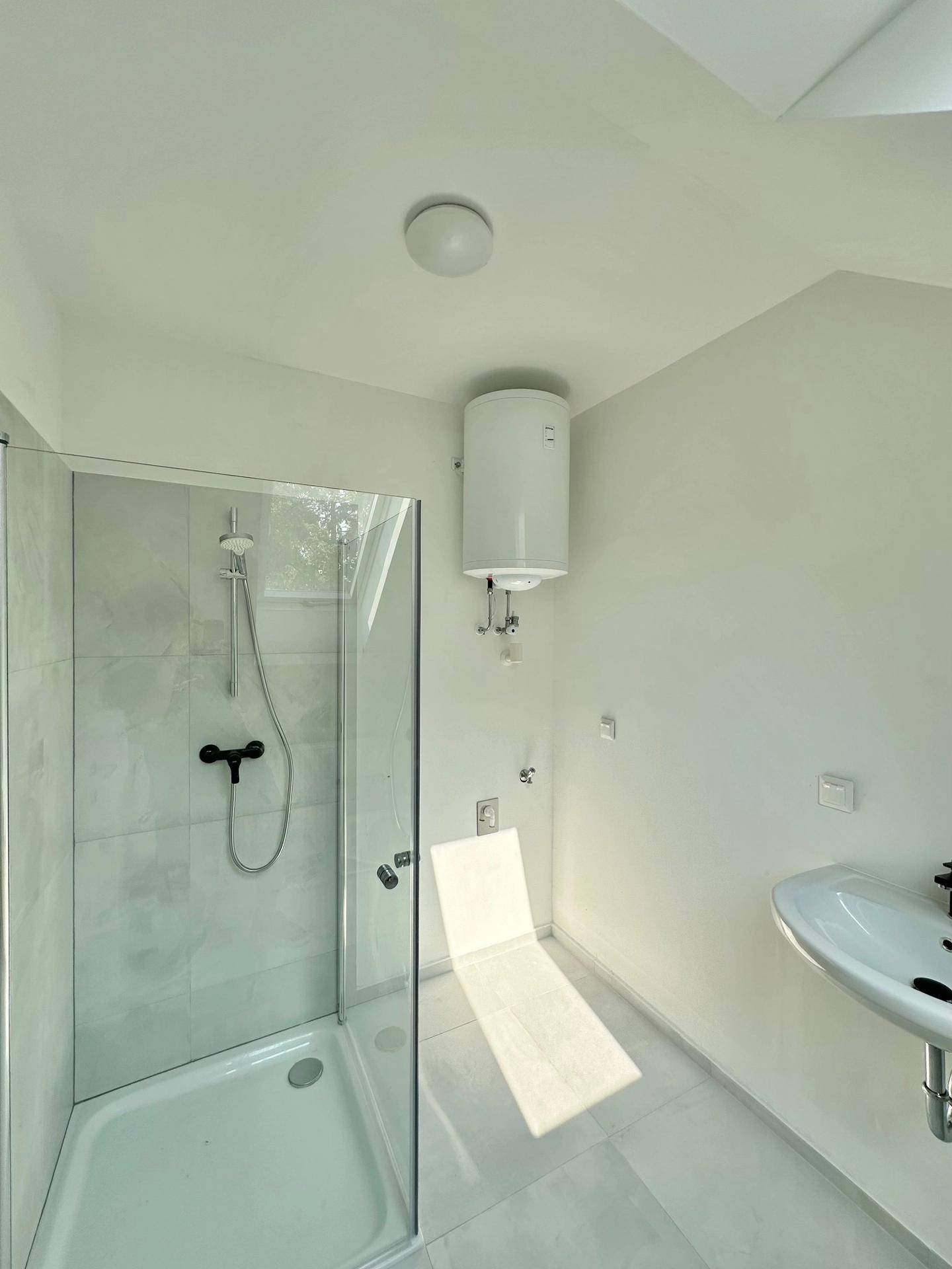 Modernes Badezimmer mit Duschkabine, Waschbecken und Boiler, hell gefliest und mit Dachfenster.