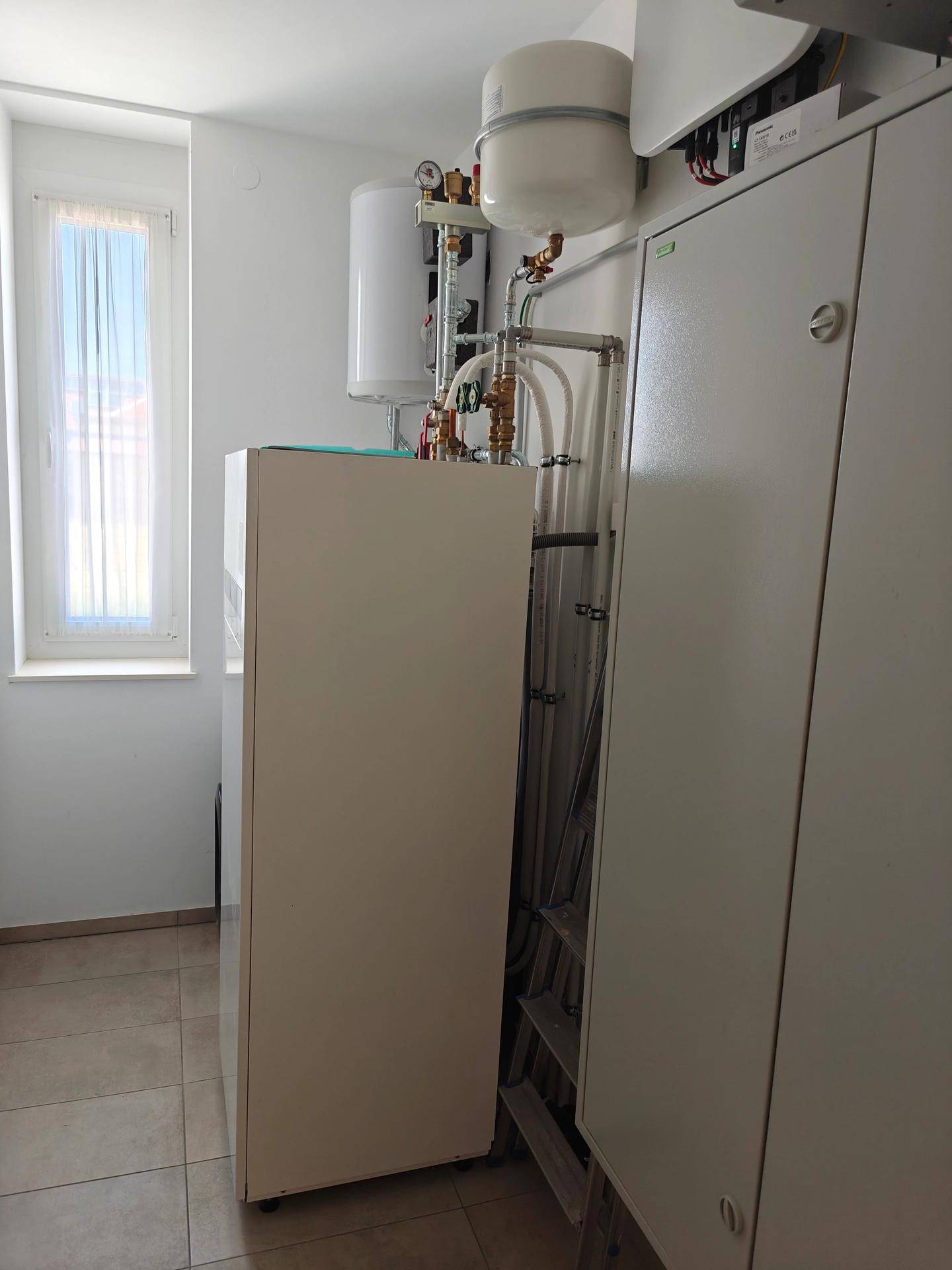 Technikraum mit Heizungsanlage, Warmwasserspeicher und Fenster für natürliche Belüftung.