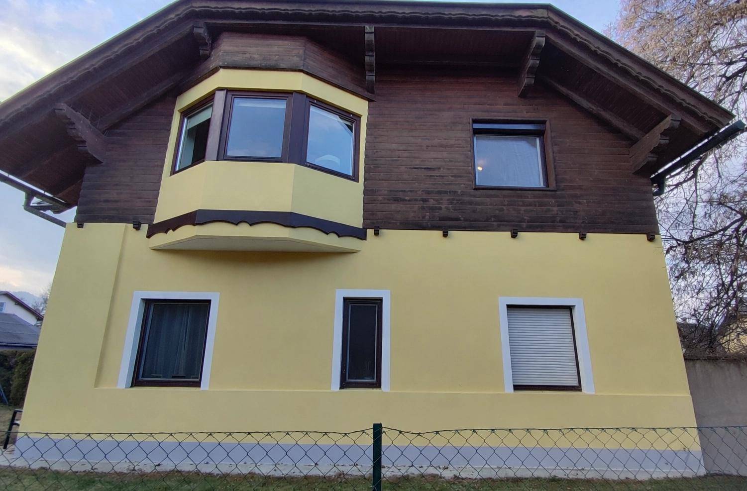 Vorderansicht des Hauses mit gelber Fassade, Holzbalkon und einem markanten Erkerfenster.