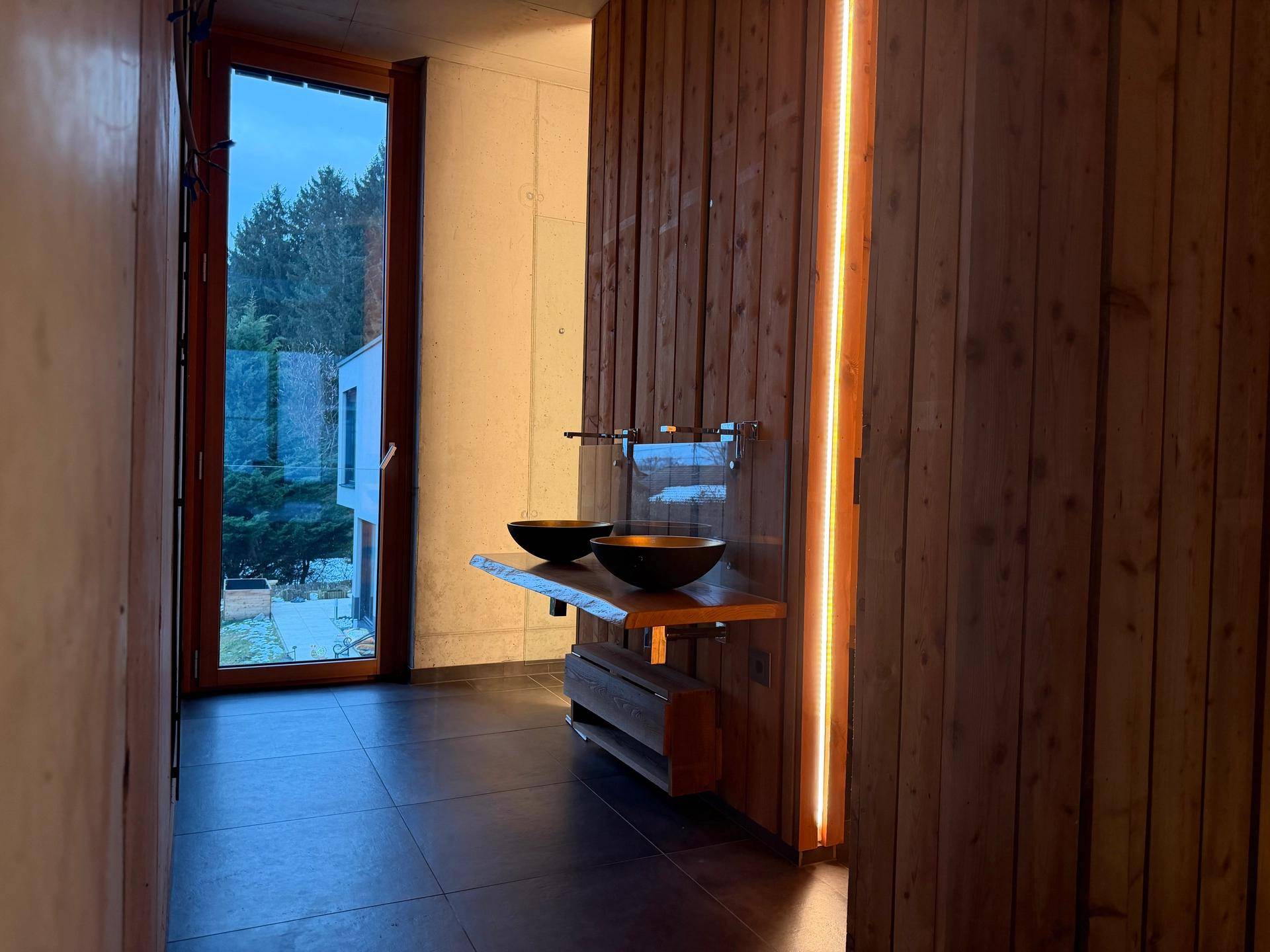 Stilvolles Badezimmer mit Holzwänden, dunklen Fliesen, goldenem Waschbecken und einem Fenster.
