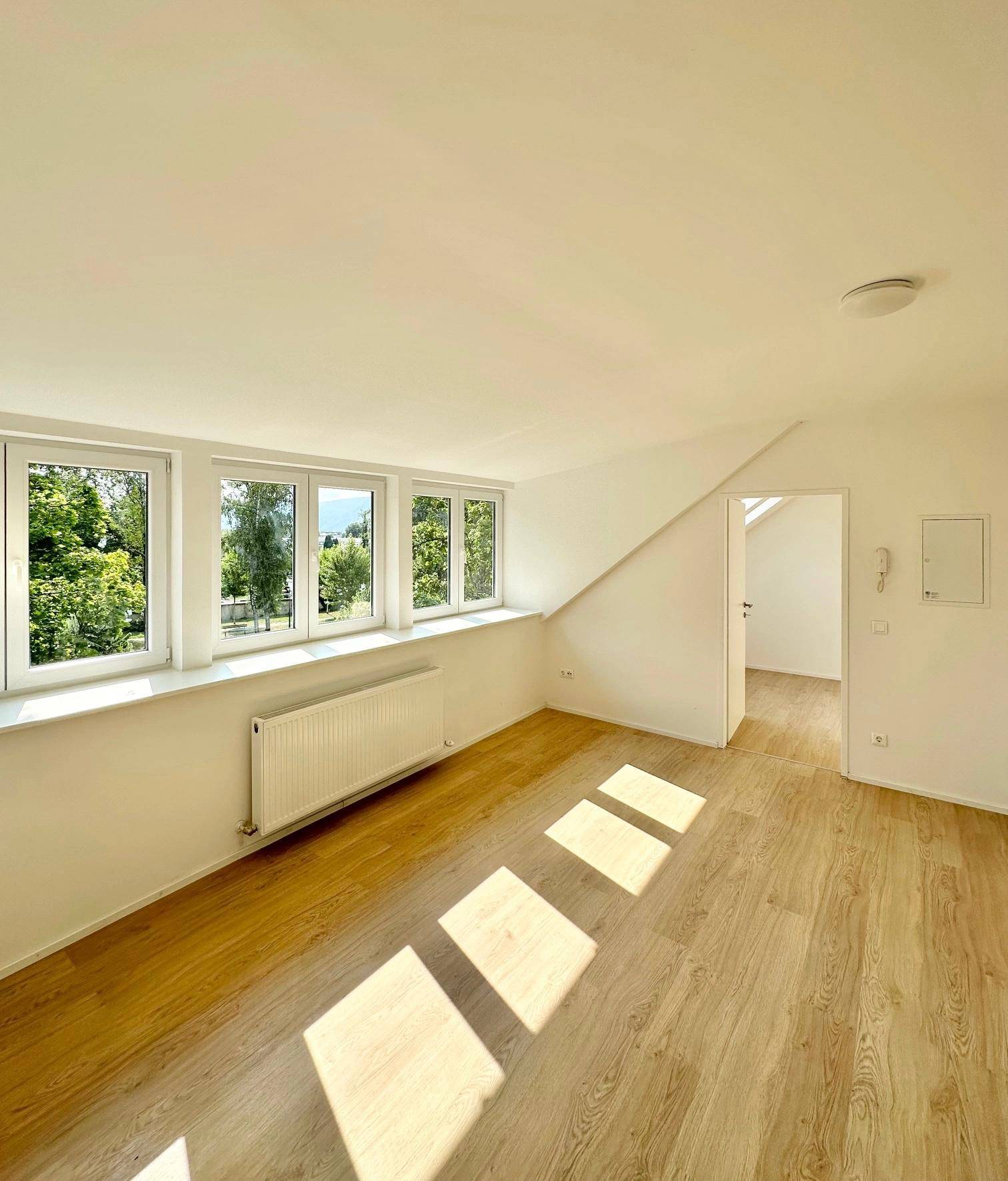 Heller Wohnraum im Dachgeschoss mit Laminatboden, Heizkörper und Blick ins Grüne durch mehrere Fenster.