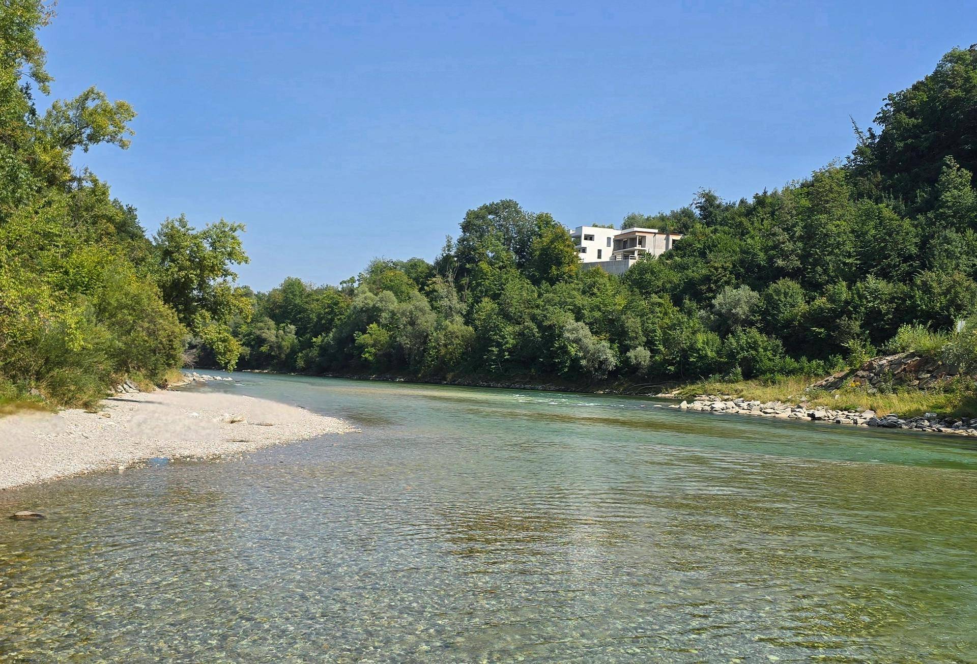 Malerische Flusslandschaft mit klarem Wasser und üppiger Vegetation, im Hintergrund ein modernes Gebäude.