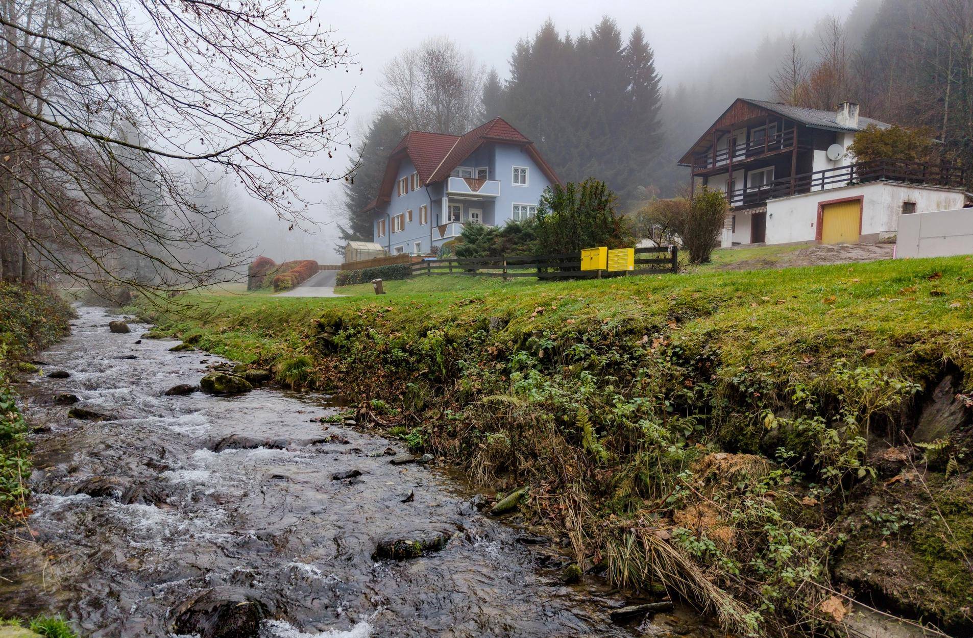 Bachlauf in einer grünen Landschaft mit Häusern und Wald im Hintergrund.