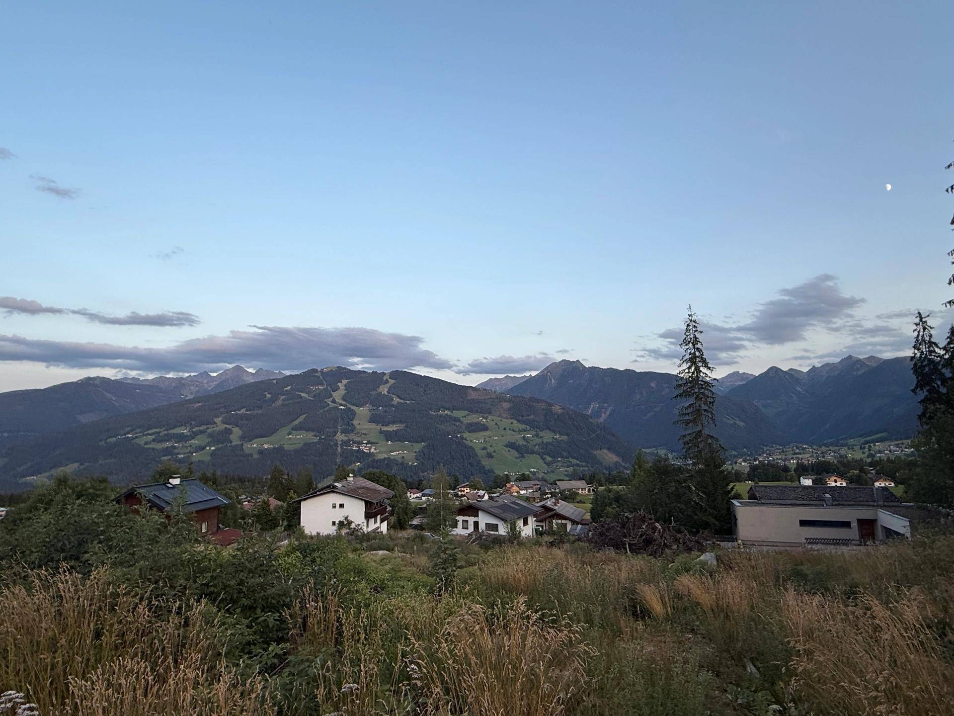 Weitläufiger Blick über die alpine Landschaft mit Häusern und bewaldeten Bergen bei Dämmerung.