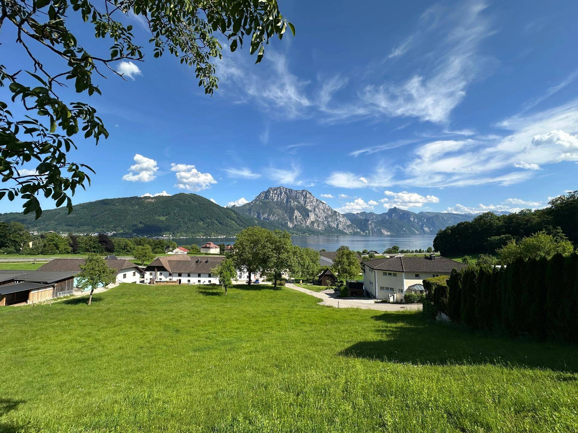 Idyllische Landschaft mit weiten Wiesen, einem See und Bergen im Hintergrund unter strahlendem Himmel.