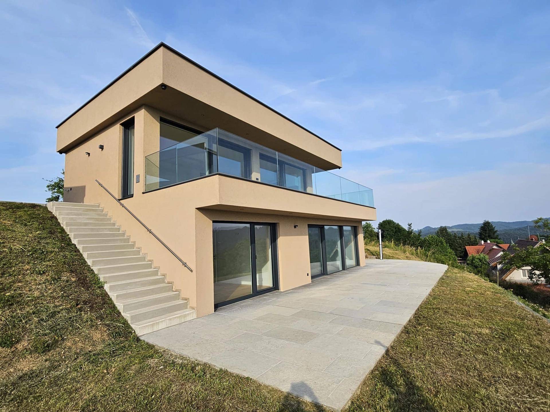 Modernes Haus mit großer Terrasse und Außentreppe, eingebettet in eine grüne Hügellandschaft.