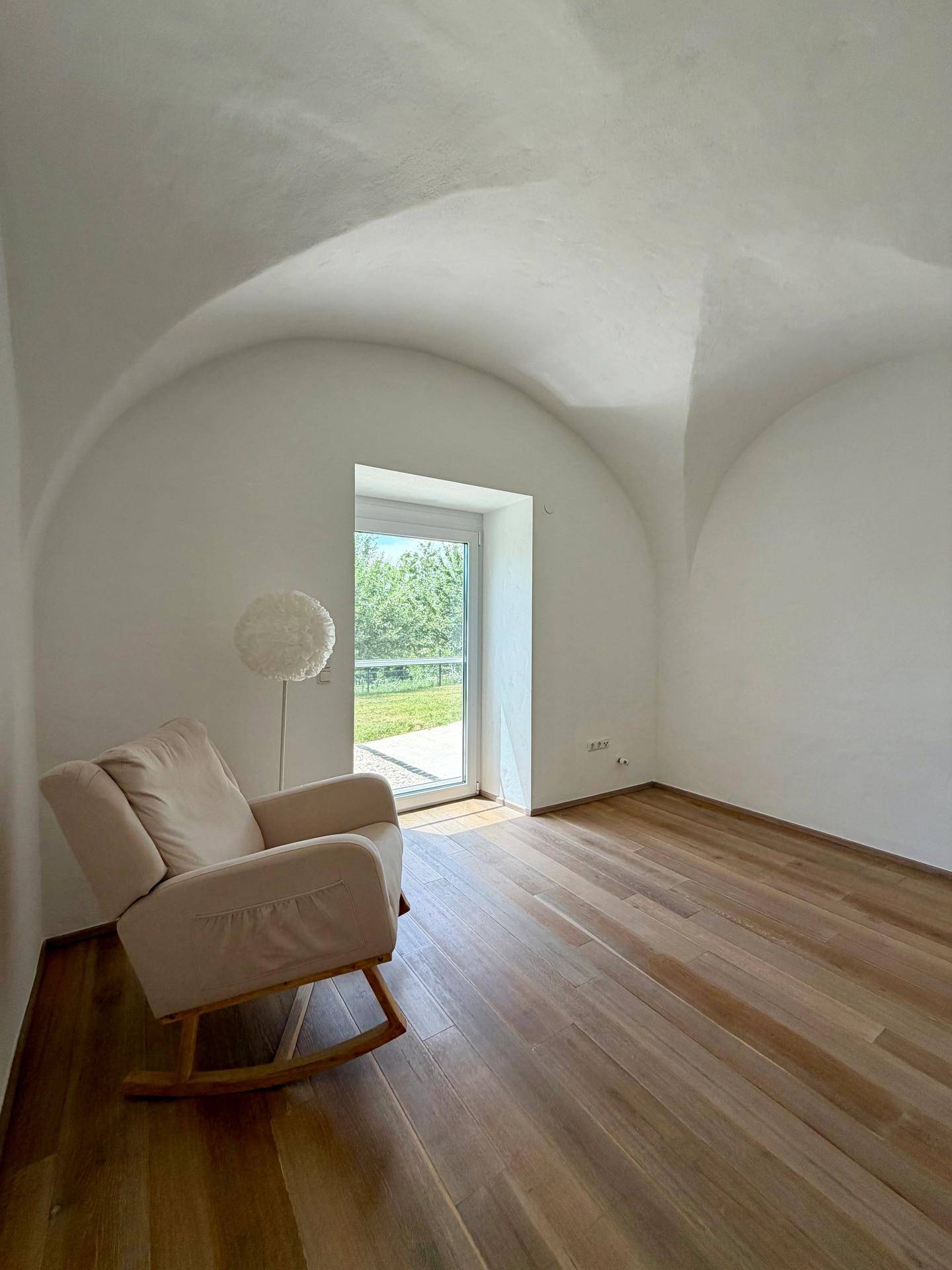Helles Zimmer mit Holzboden, Gewölbedecke und einem bequemen Sessel am Fenster.