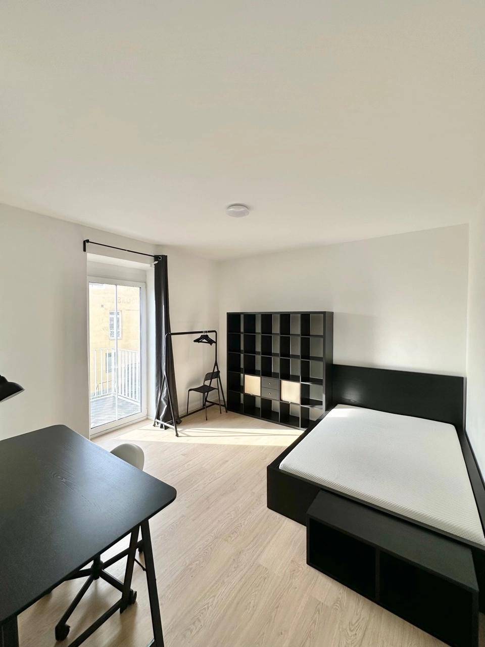 Helles Schlafzimmer mit modernem Bett, Schreibtisch und Regal, sowie Zugang zu einem französischen Balkon.