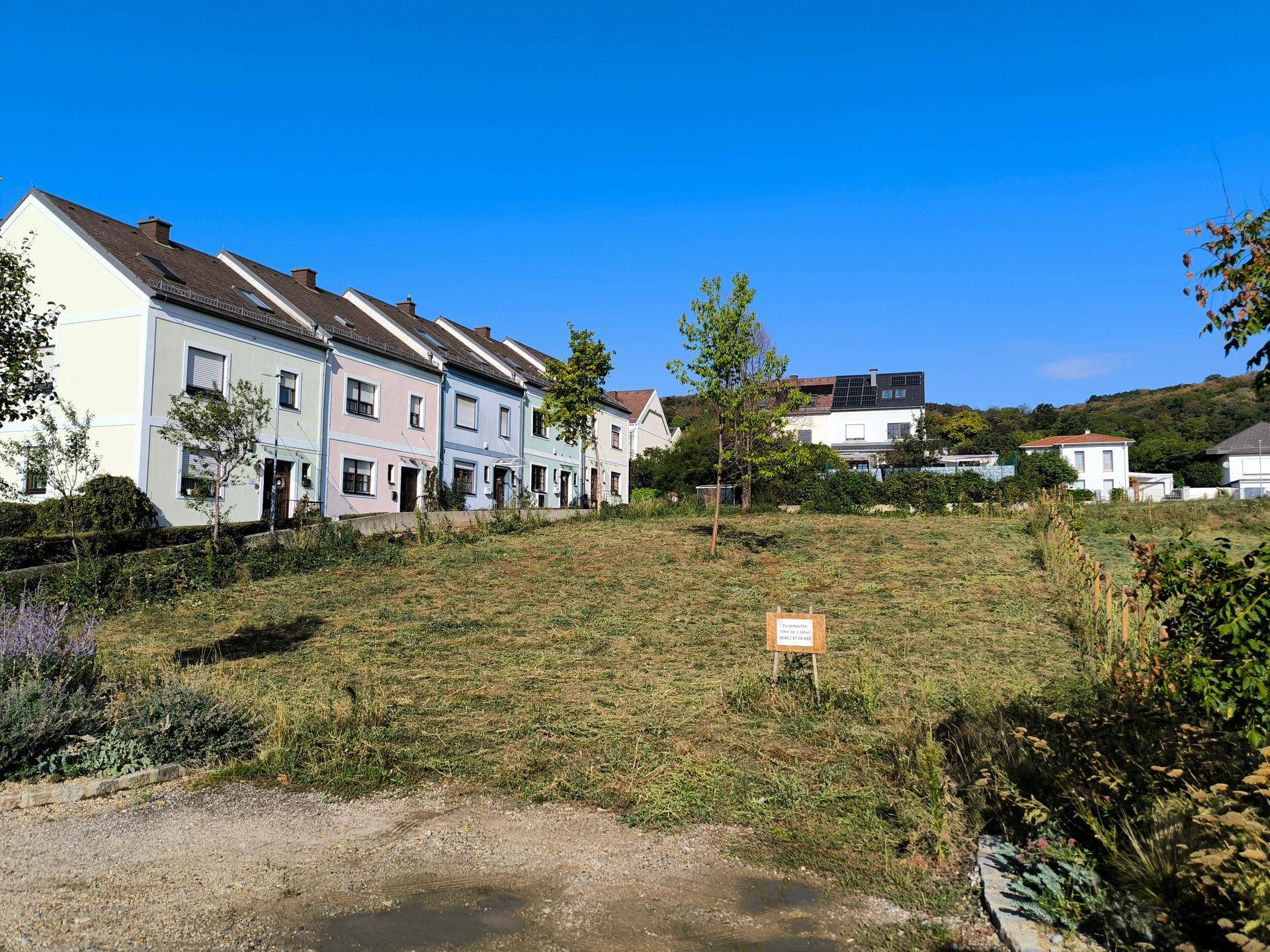 Grünes Baugrundstück mit Blick auf eine Reihe farbiger Wohnhäuser und einen klaren blauen Himmel.