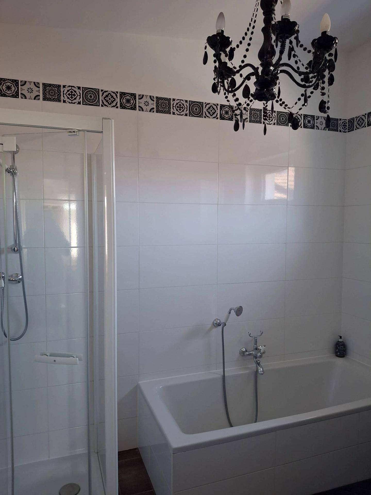 Helles Badezimmer mit weißen Fliesen, Badewanne, Duschkabine und einem dekorativen Kronleuchter.
