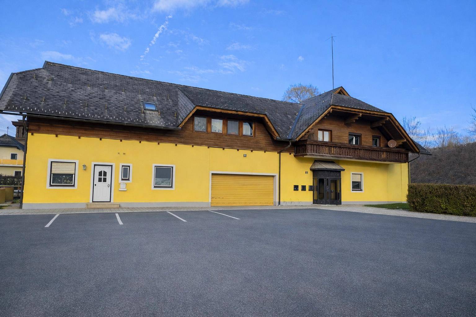 Gelbes Gebäude mit Holzverkleidung, Satteldach und großem Parkplatz davor unter blauem Himmel.