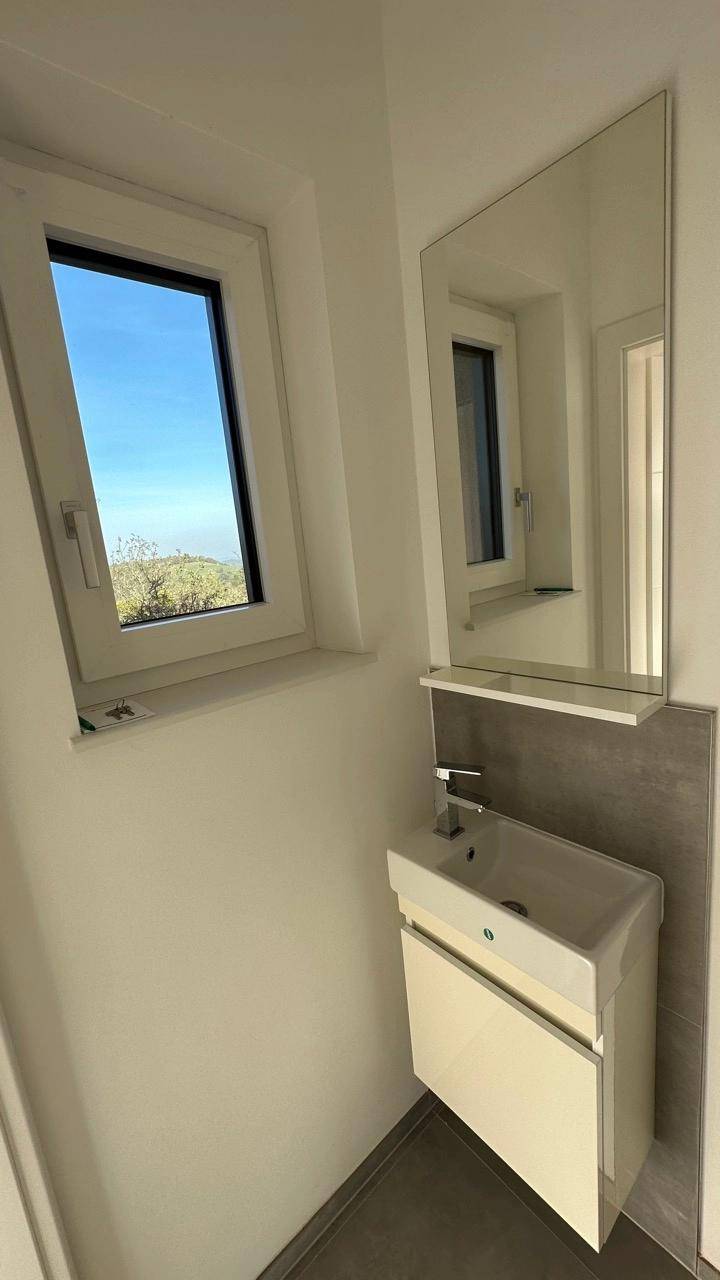 Modernes Gäste-WC mit kleinem Waschbecken, Spiegel und Fenster, das viel Tageslicht hereinlässt.