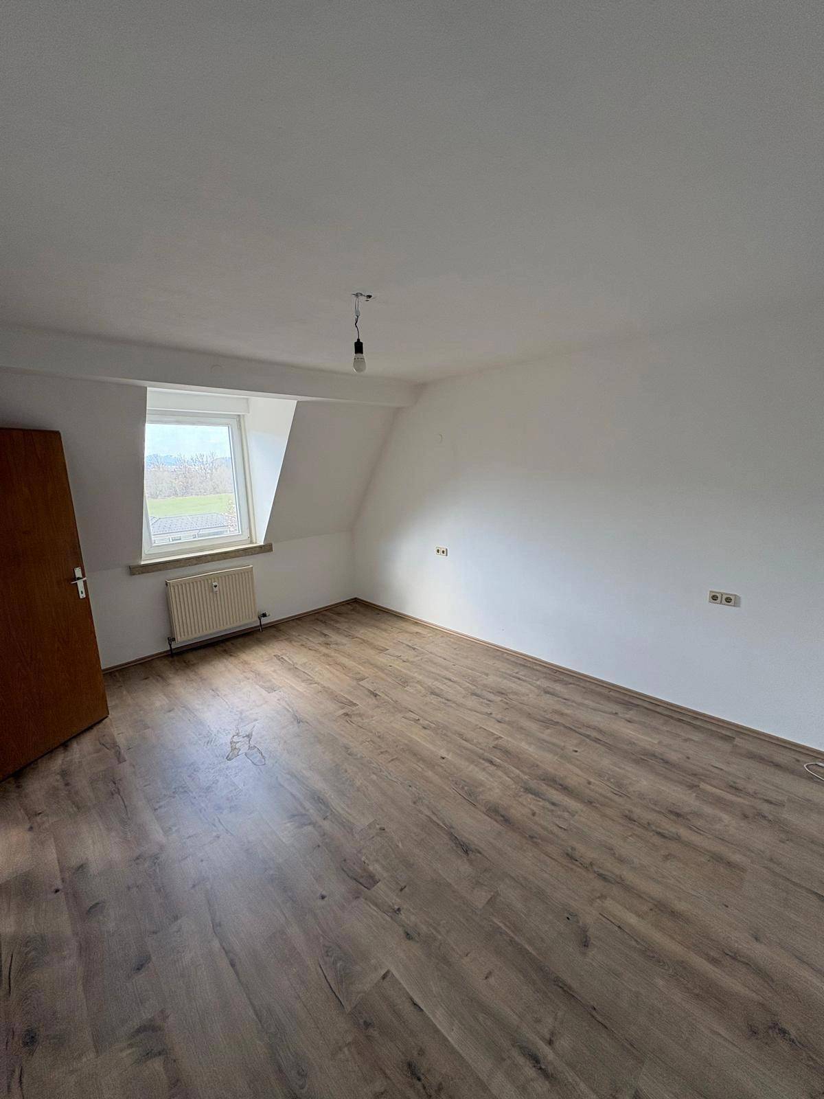 Leeres Zimmer mit Laminatboden, Dachschräge und einem Fenster, das viel natürliches Licht hereinlässt.