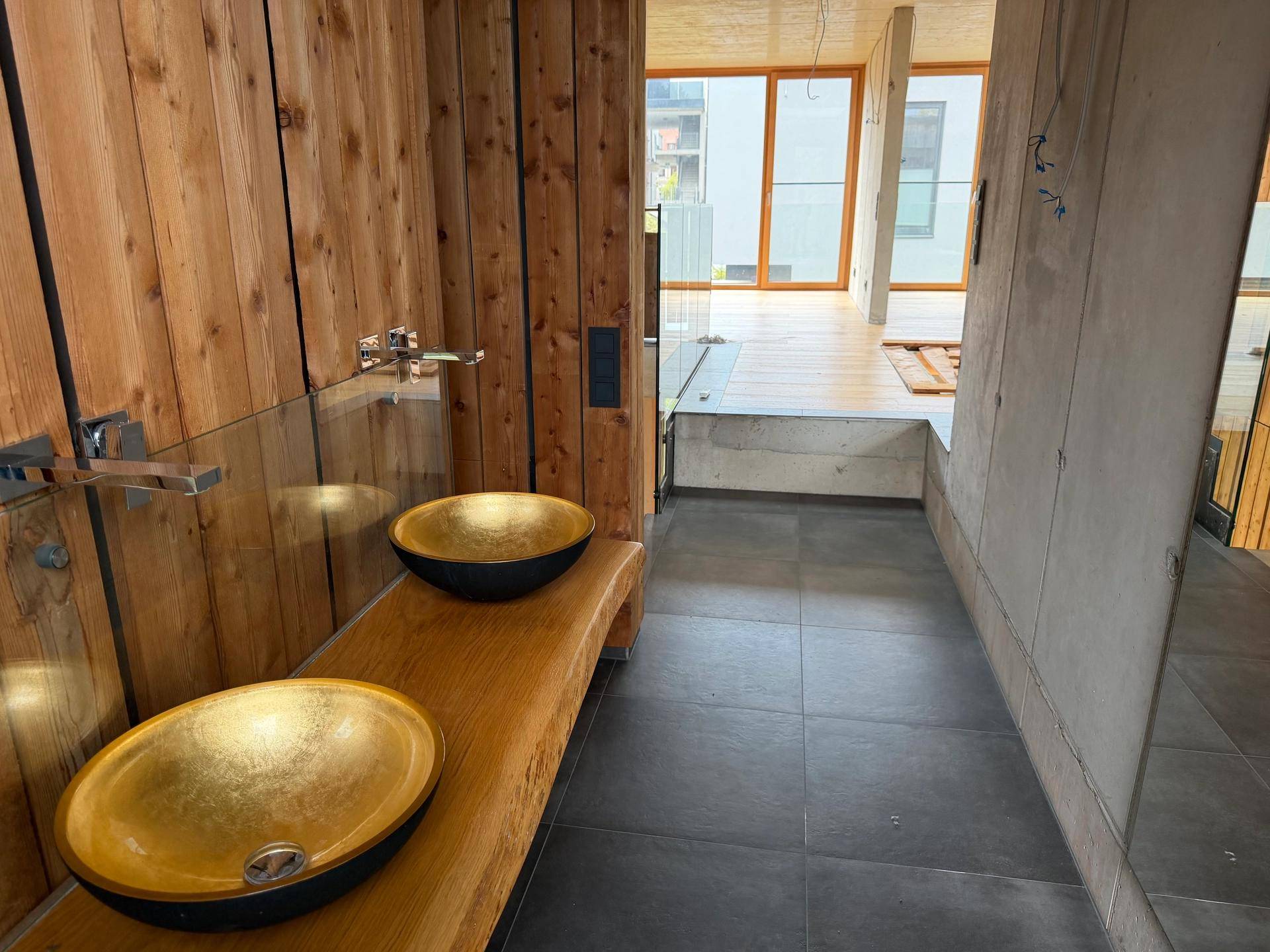 Modernes Badezimmer mit Holzwänden, dunklen Fliesen und zwei einzigartigen goldenen Aufsatzwaschbecken.