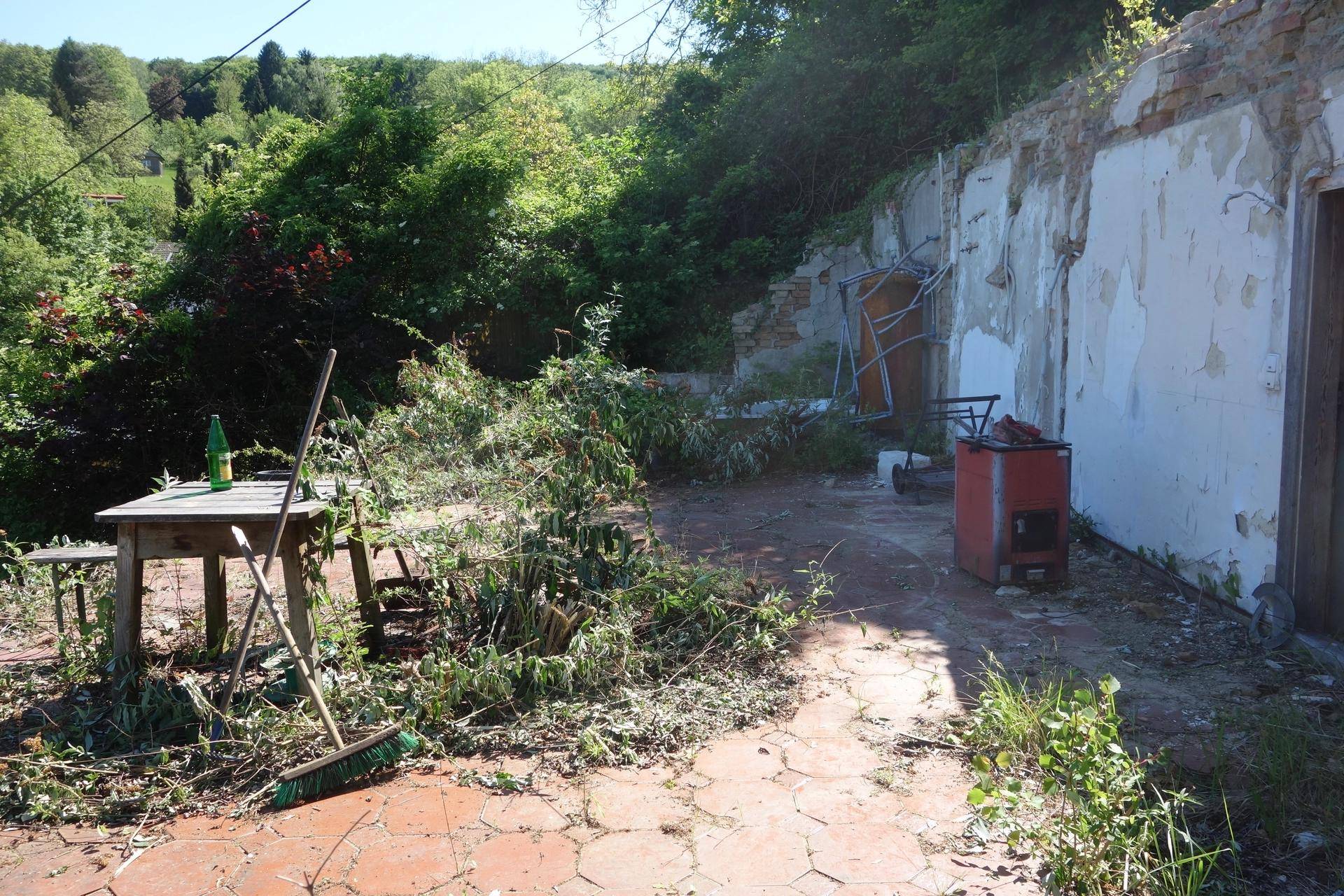 Ungepflegte Terrasse mit altem Holztisch und Blick auf die bewachsene Umgebung und eine Ruine.