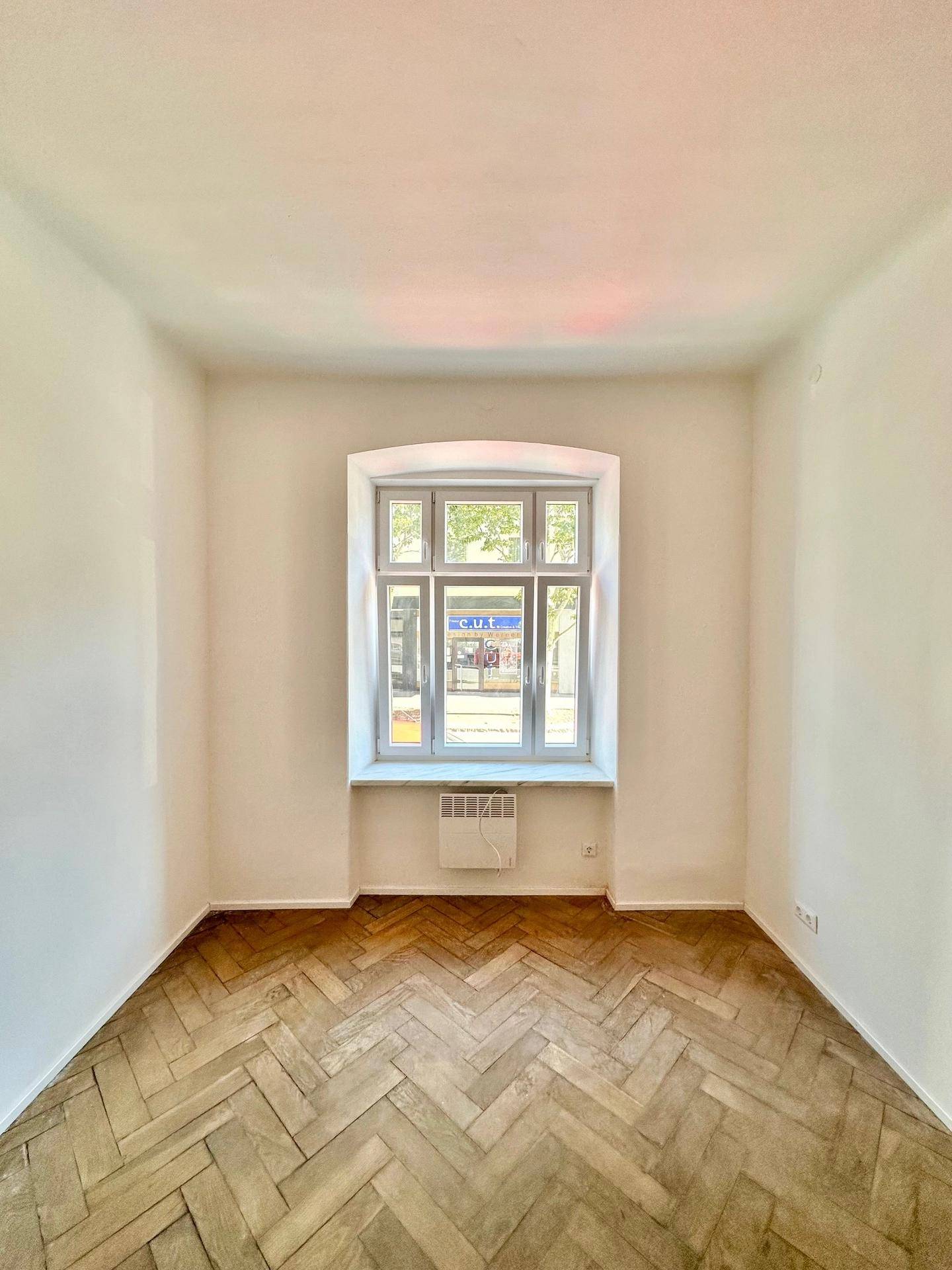 Heller Raum mit Fischgrätparkett und großem Fenster zur Straße.