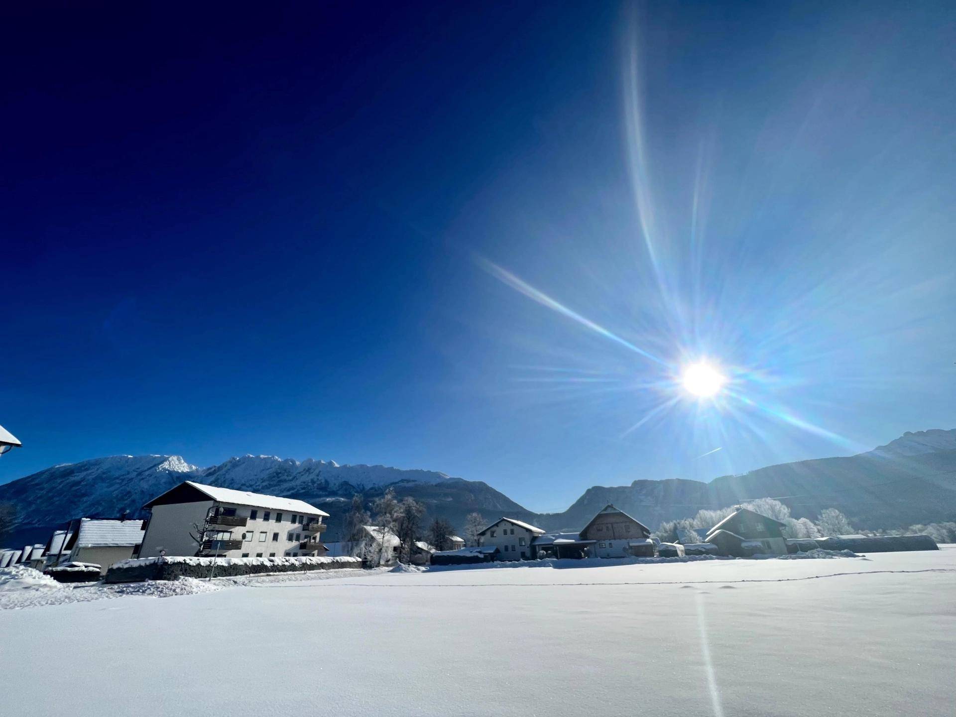 Sonnige Winterlandschaft mit verschneiten Häusern und Feldern vor einer beeindruckenden Bergkette.