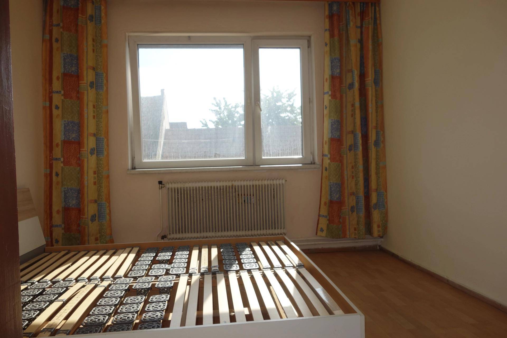 Leeres Schlafzimmer mit hellem Laminatboden, Fenster und bunten Vorhängen.