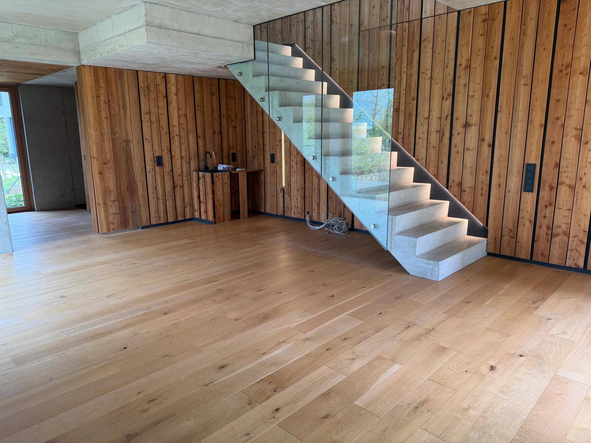 Offener Wohnbereich mit Holzwänden, Sichtbetondecke und einer modernen Treppe mit Glasgeländer.