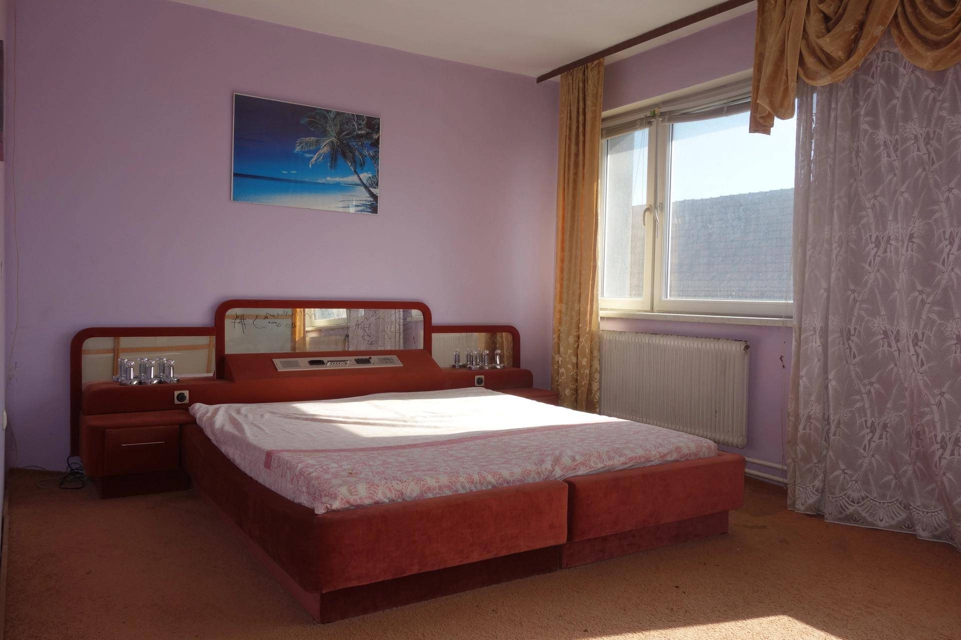 Schlafzimmer mit lila Wänden, Teppichboden, rotem Bett und einem Fenster mit Vorhängen.