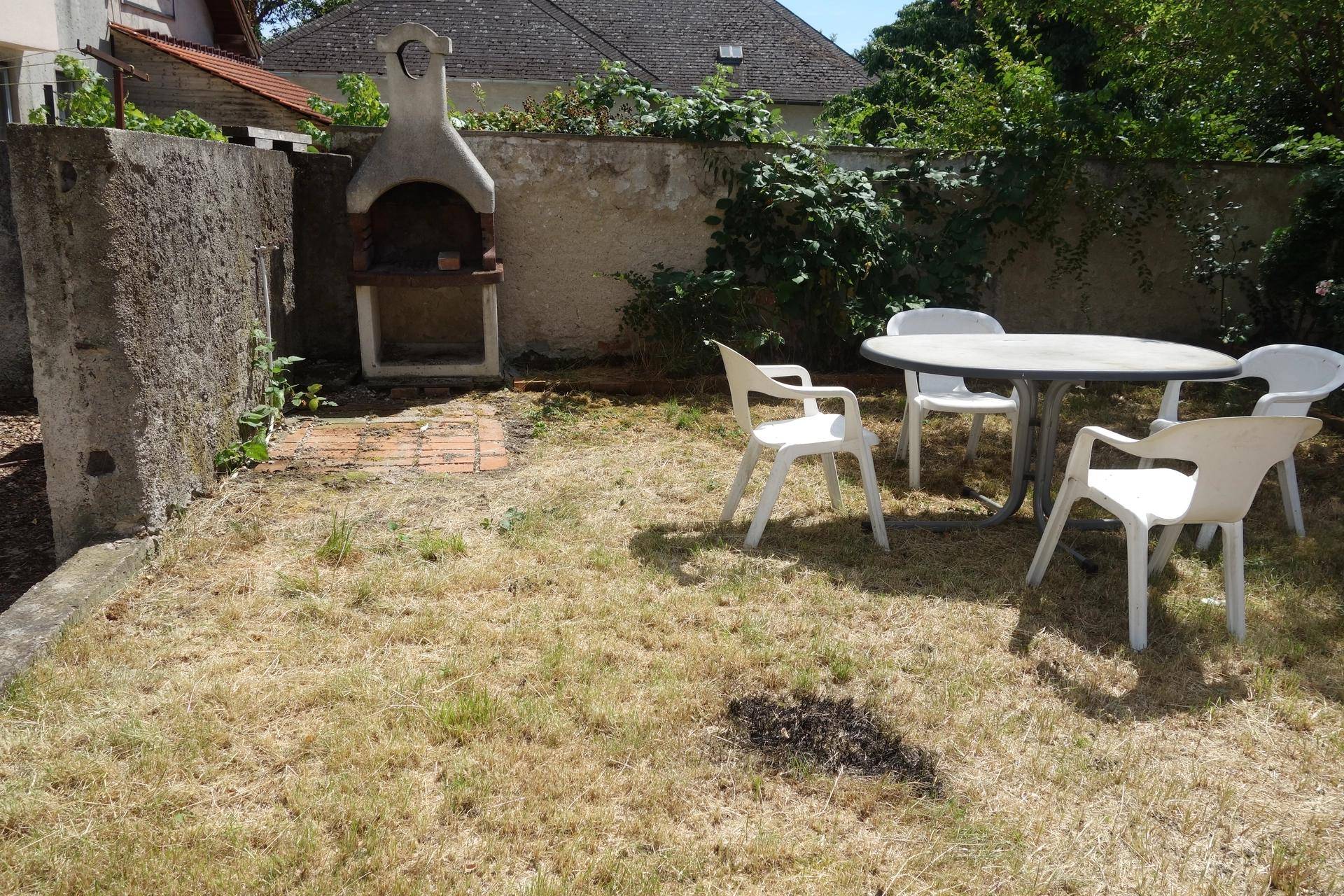 Kleiner Gartenbereich mit gemauertem Grill und einfachen Gartenmöbeln auf trockenem Rasen.