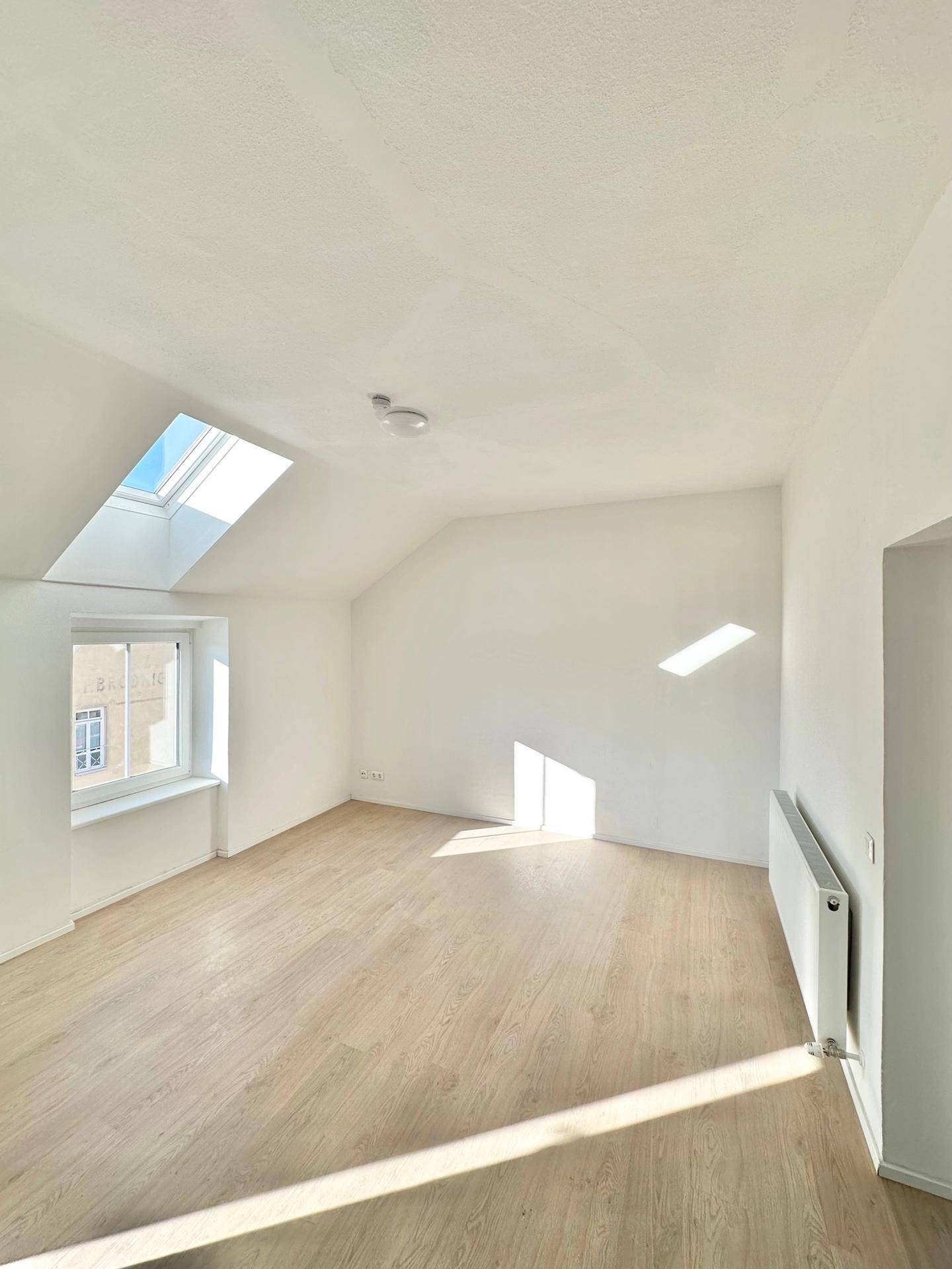 Lichtdurchfluteter Wohnraum im Dachgeschoss mit Laminatboden, Heizkörper und einem großen Dachfenster.