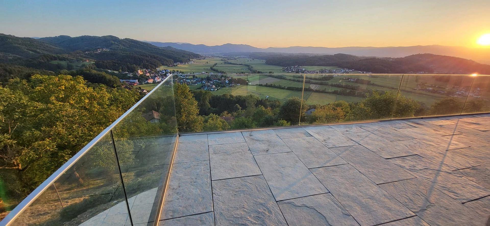 Geräumiger Balkon mit Steinplatten und Glasgeländer, bietet einen weiten Blick über die grüne Landschaft.