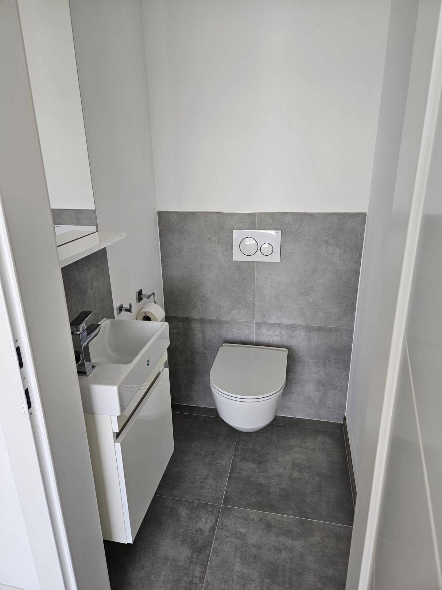 Kompaktes Gäste-WC mit wandhängender Toilette, kleinem Waschbecken und modernen grauen Fliesen.