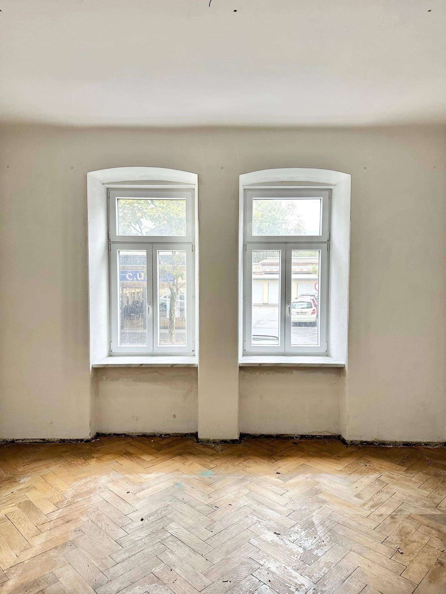 Leerer Raum mit Fischgrätparkett und zwei Fenstern mit Blick auf die Straße.