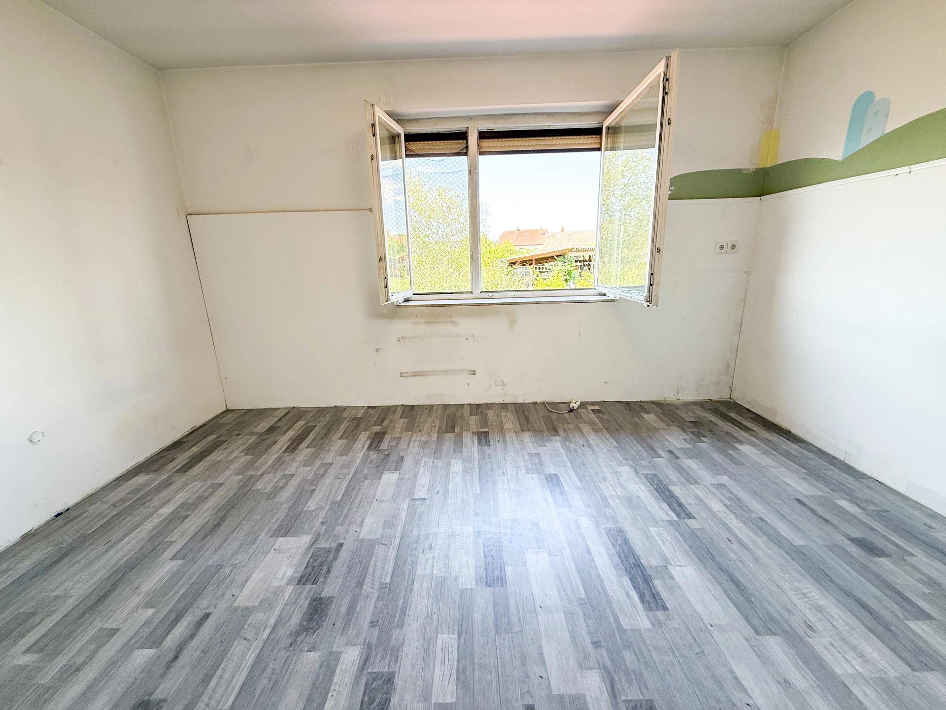 Leeres Zimmer mit grauem Laminatboden und Fenster, teilweise mit bunter Wanddekoration.