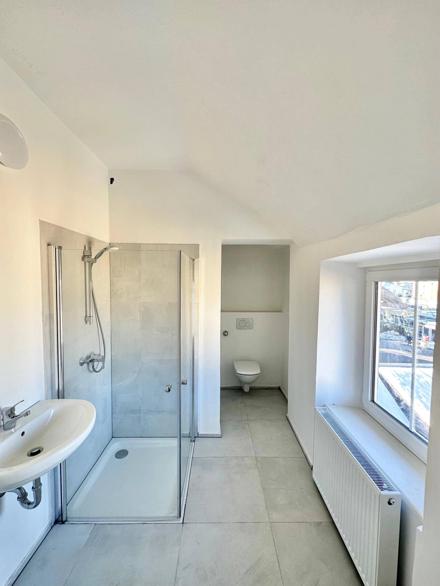 Modernes Badezimmer mit Duschkabine, Waschbecken und separatem WC-Bereich, hell gefliest und mit Fenster.