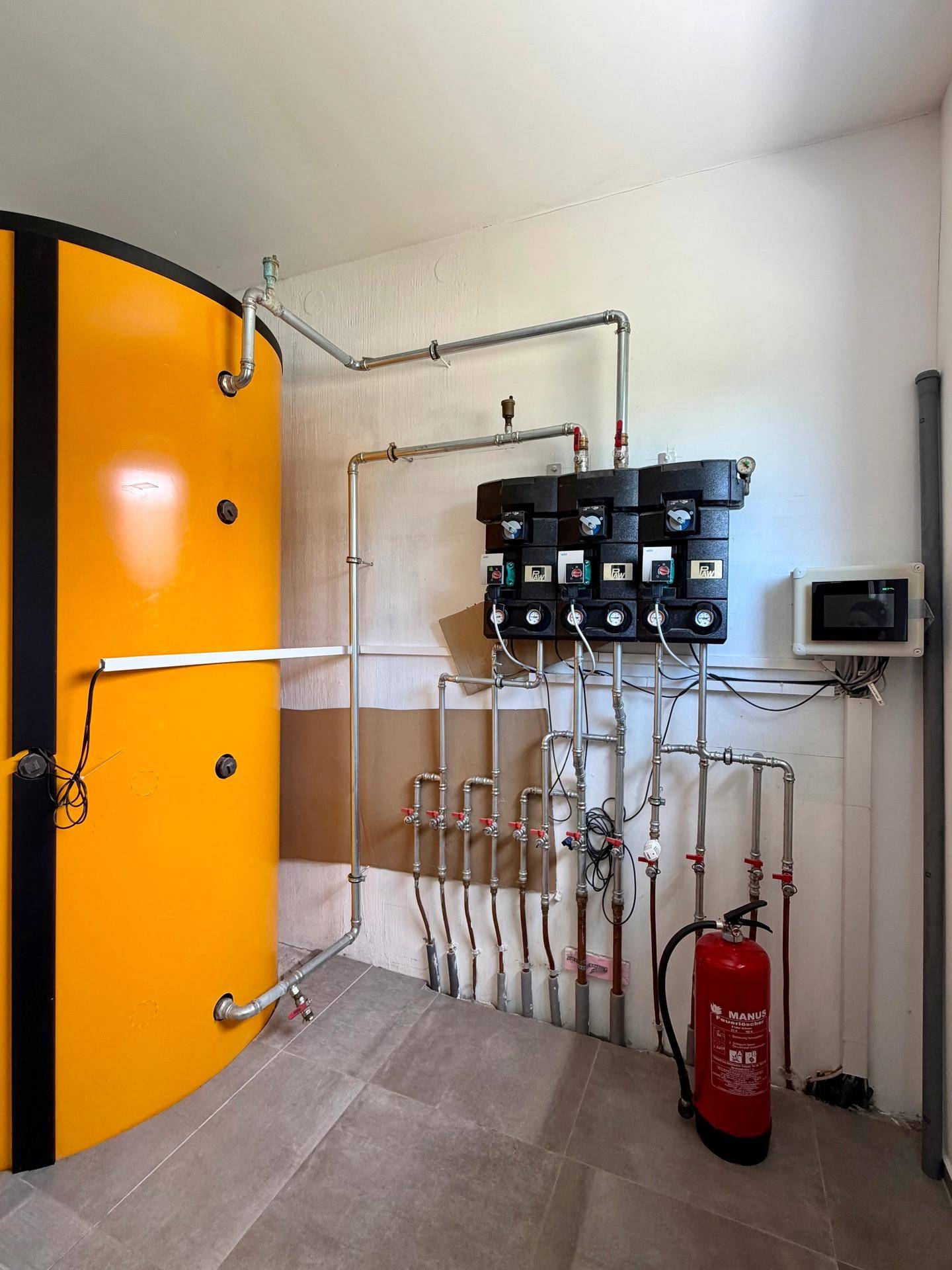 Technikraum mit großem Warmwasserspeicher, Heizungsanlage und sichtbaren Rohrleitungen.