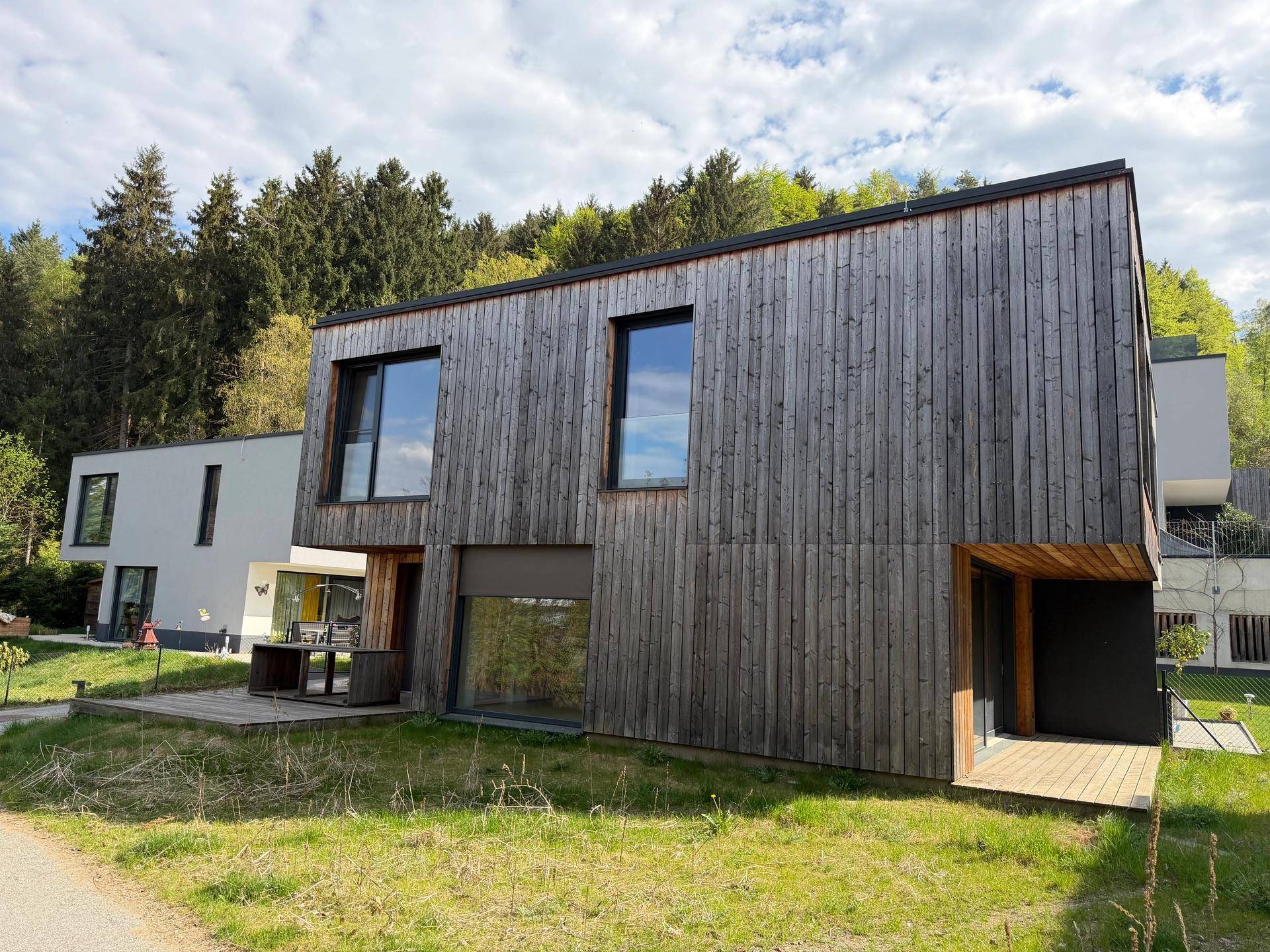 Ansicht eines modernen Hauses mit vertikaler Holzverkleidung und großen Fensterflächen in ländlicher Umgebung.