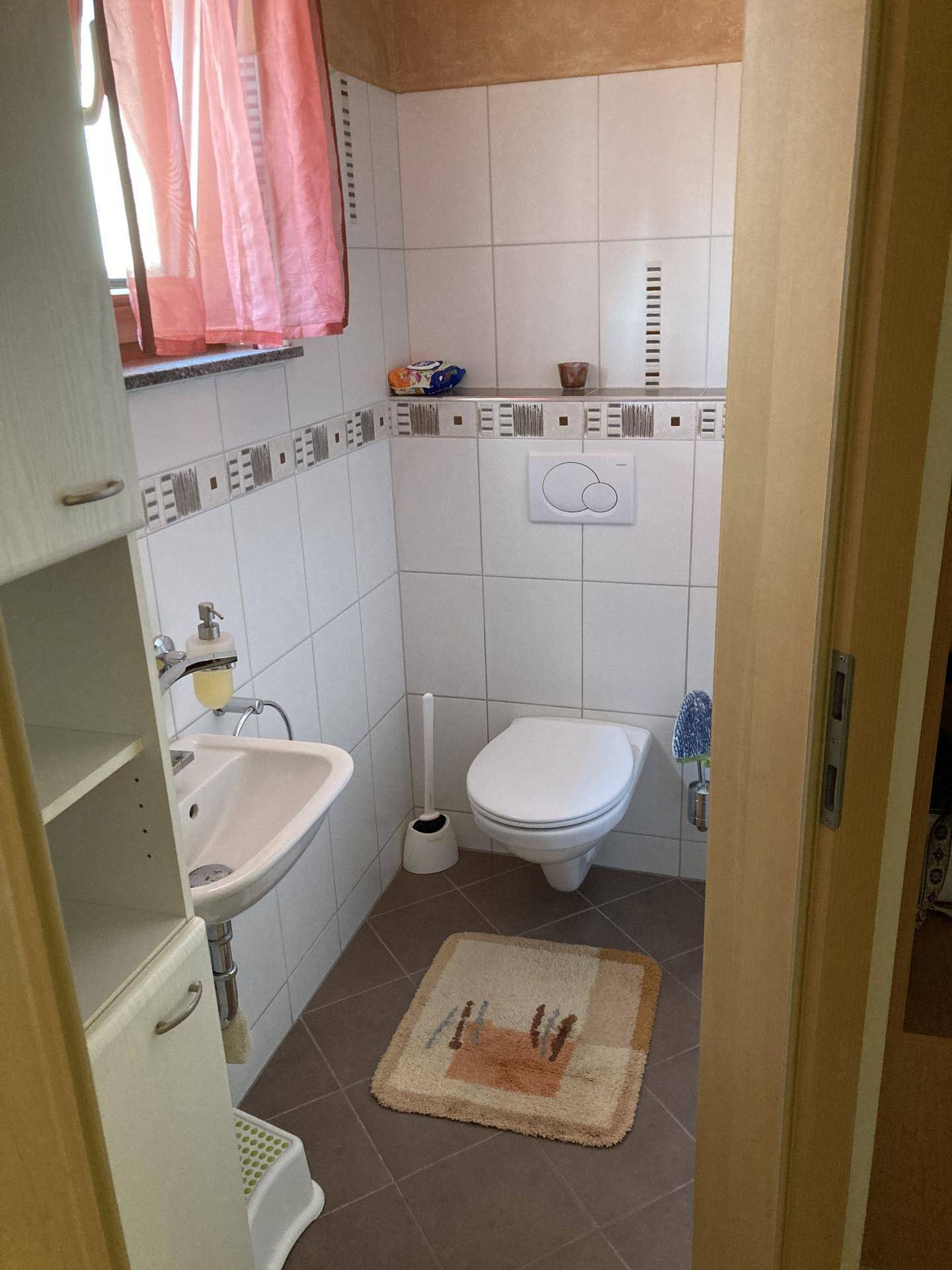 Kleines Gäste-WC mit Fenster, Wand-WC und Waschbecken in hellen Fliesen.